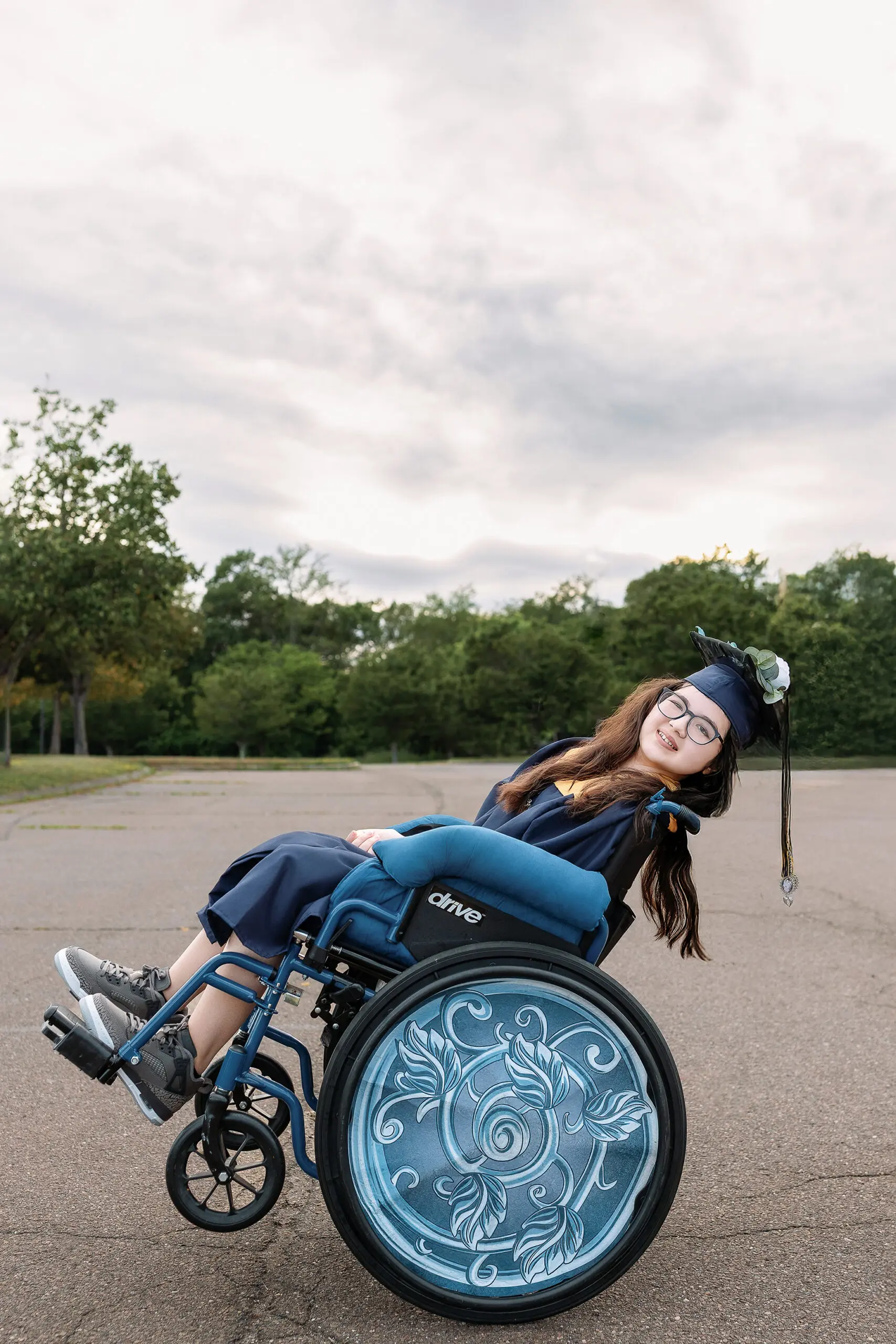 A young woman in a graduation cap and gown smiles while sitting in a wheelchair with decorative blue designs on the wheels. She is outdoors on a paved area with greenery and trees in the background.