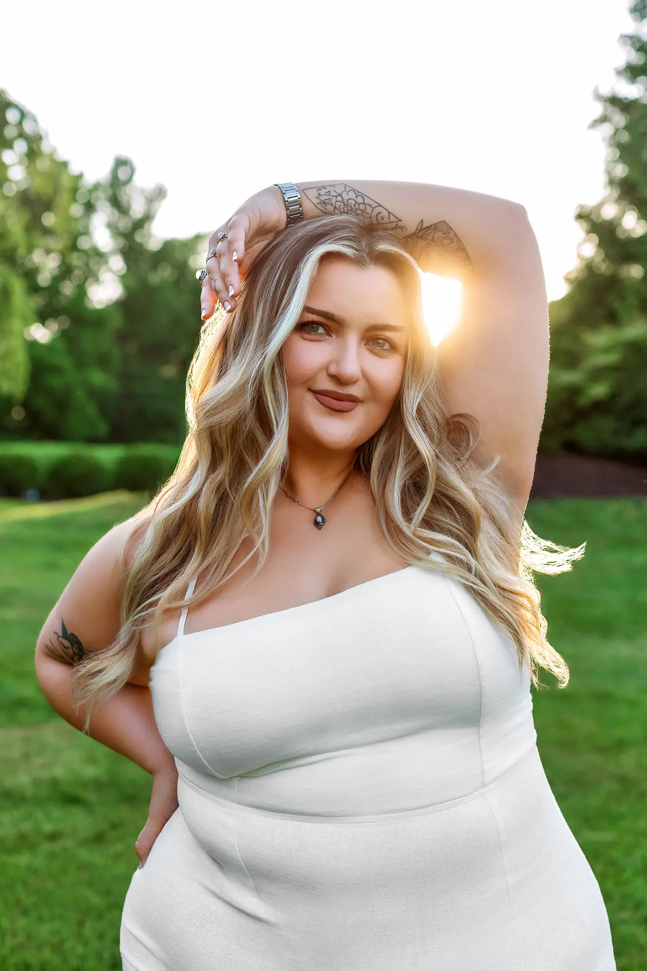 A woman in a white dress stands outdoors on green grass during her CT senior session, smiling with one hand on her hip and the other arm raised above her head. Sunlight highlights her long blonde hair and trees frame the glowing background.