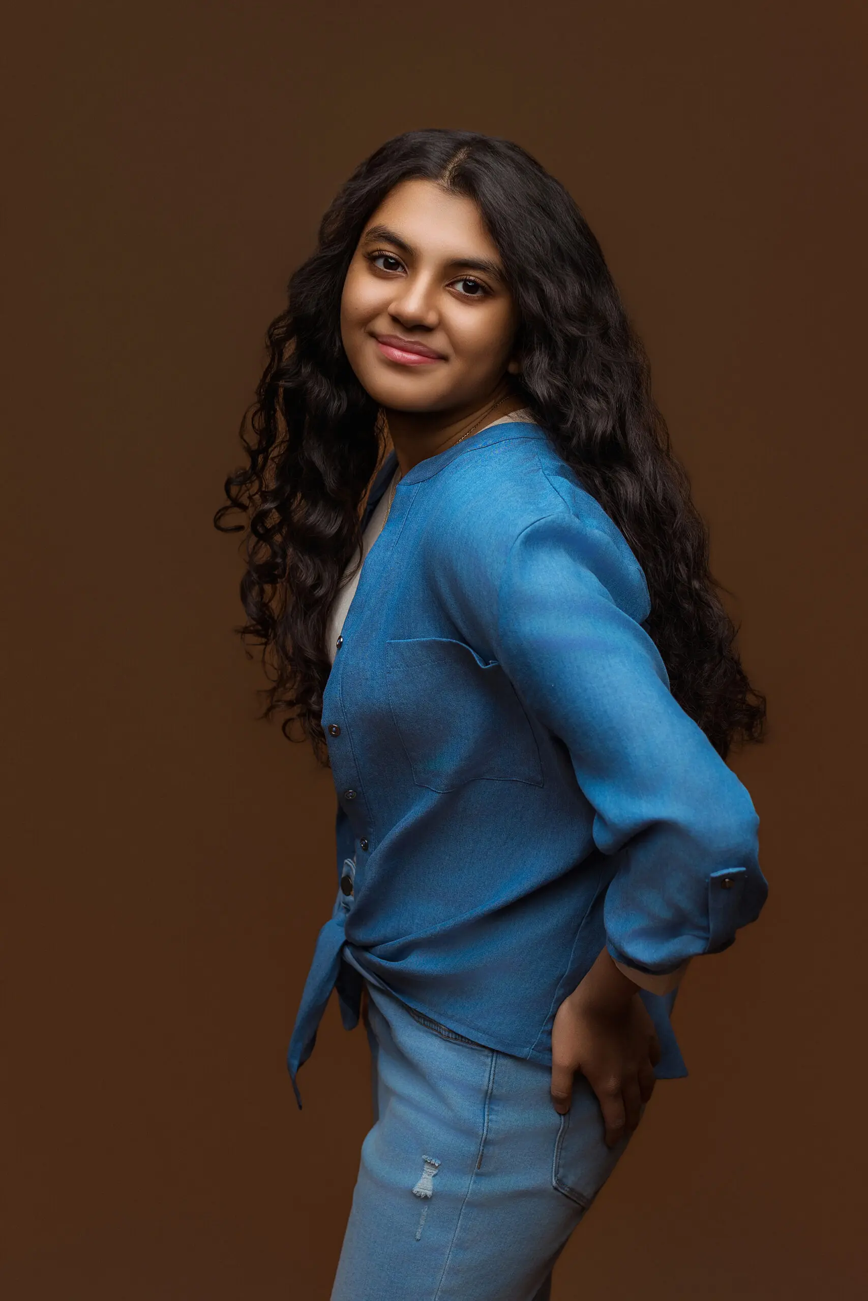 A young woman with long, wavy dark hair poses against a brown background for her CT senior photos, wearing a blue button-up shirt and light blue jeans as she smiles gently at the camera.