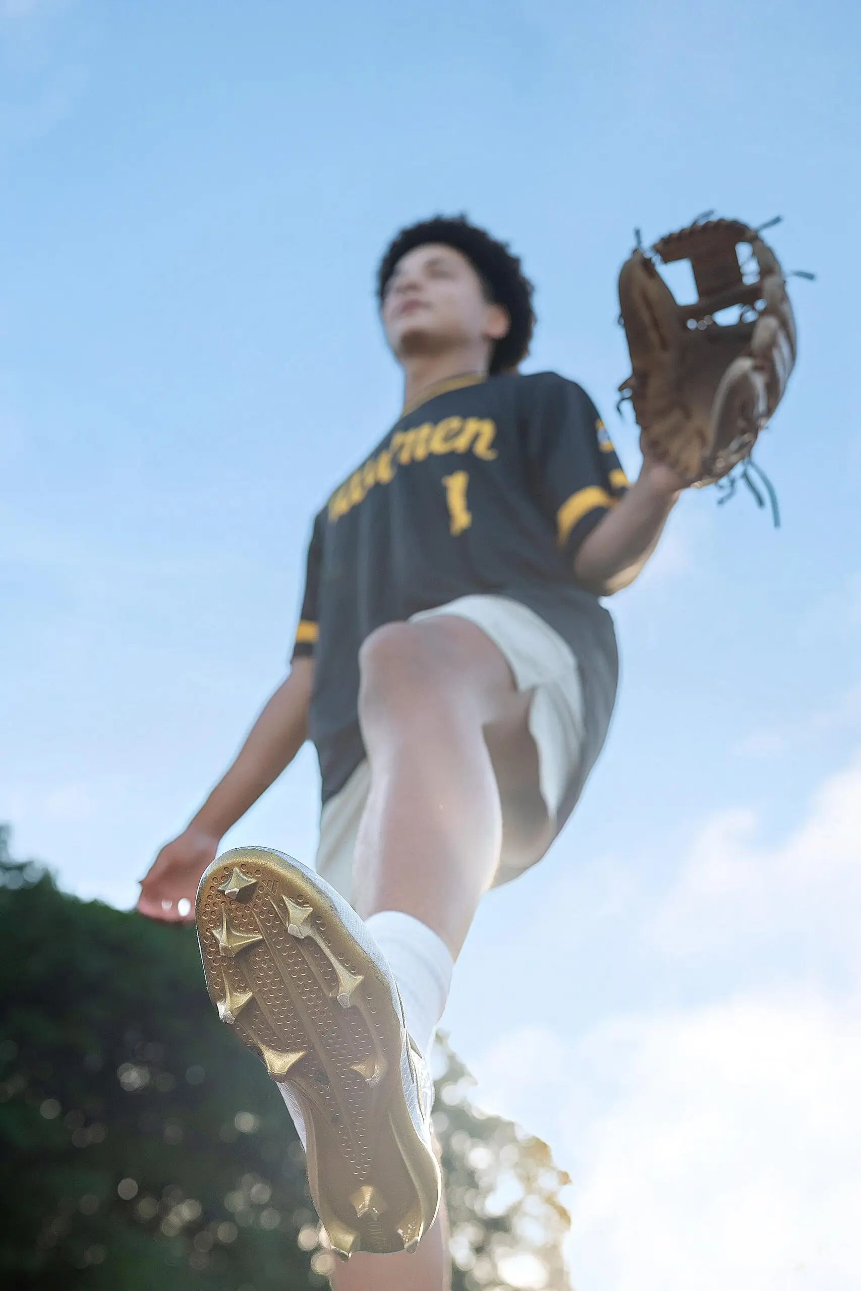 Low-angle view of a baseball player in a black and yellow jersey and glove, raising one leg with cleats visible against a blue sky—perfect inspiration for a dynamic senior portrait CT session.