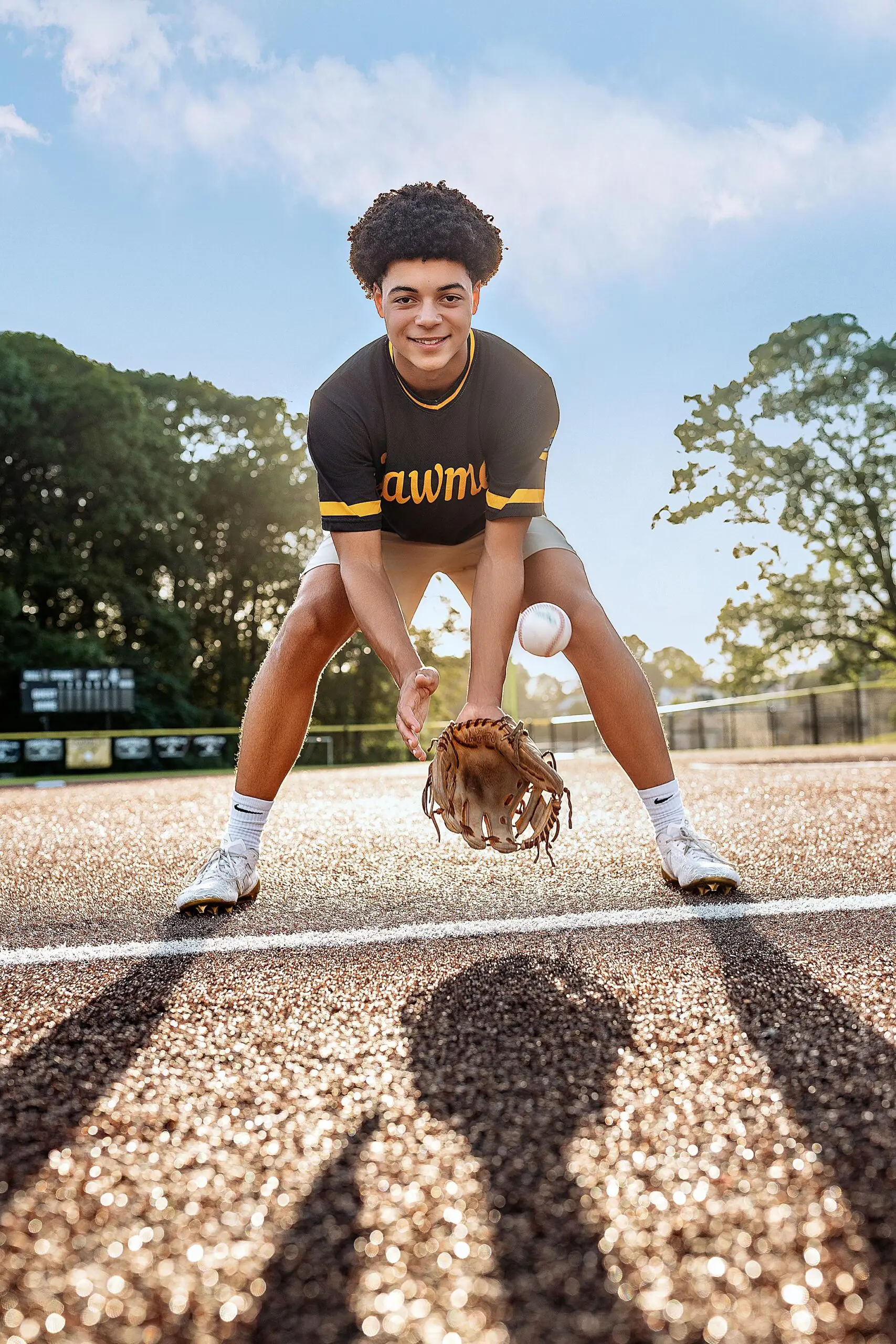 A young baseball player in uniform crouches to catch a baseball with a glove on a sunny Connecticut field, smiling at the camera under a bright sky.