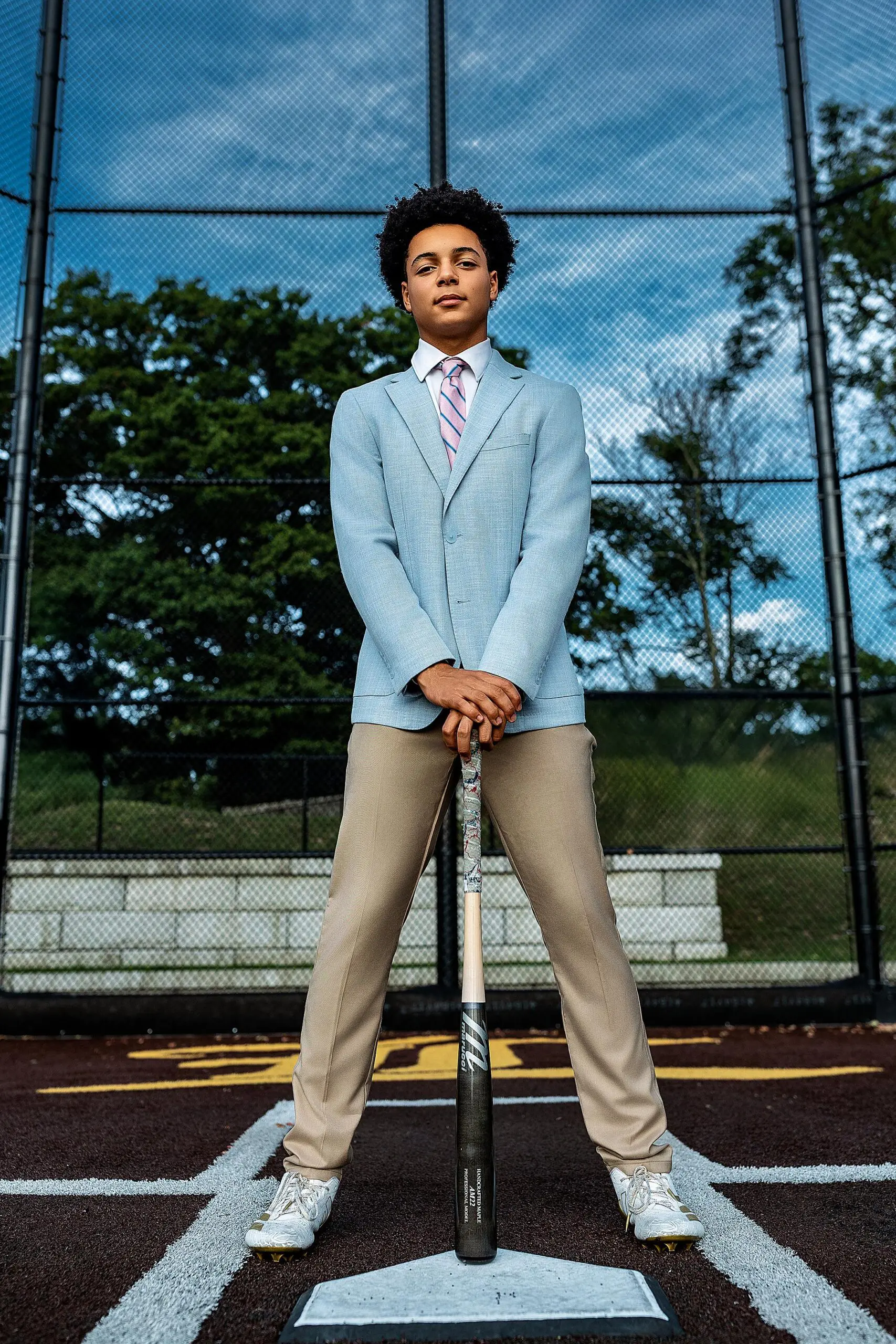 A young person in a light blue suit jacket, tie, and khaki pants stands confidently on a baseball field for their CT senior photos, holding a bat upright with both hands, against a backdrop of chain-link fence and trees.
