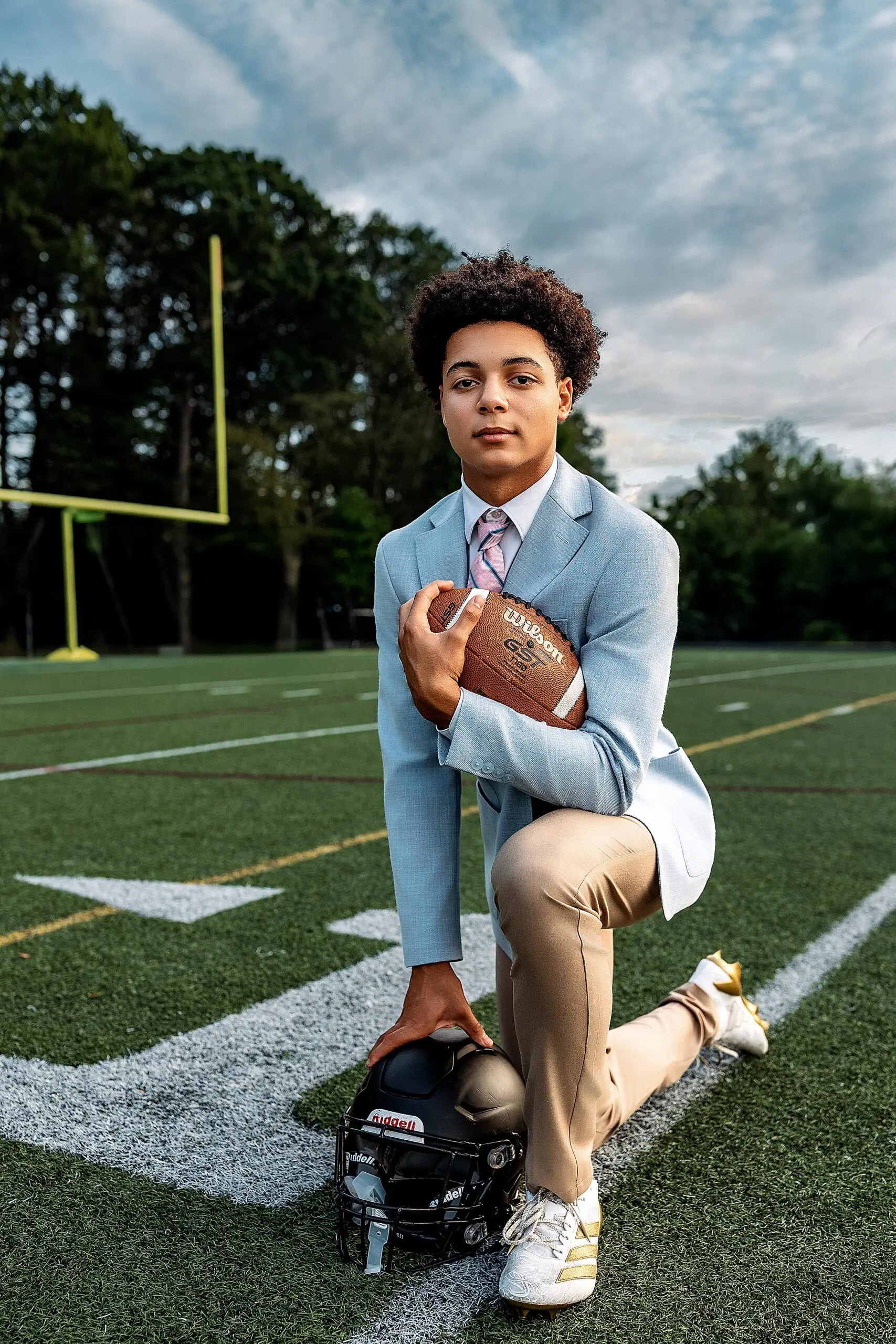 A young man wearing a light blue blazer, khaki pants, and white sneakers kneels on a football field for his CT senior portraits, holding a football and resting his arm on a black helmet, with goalposts and trees in the background.
