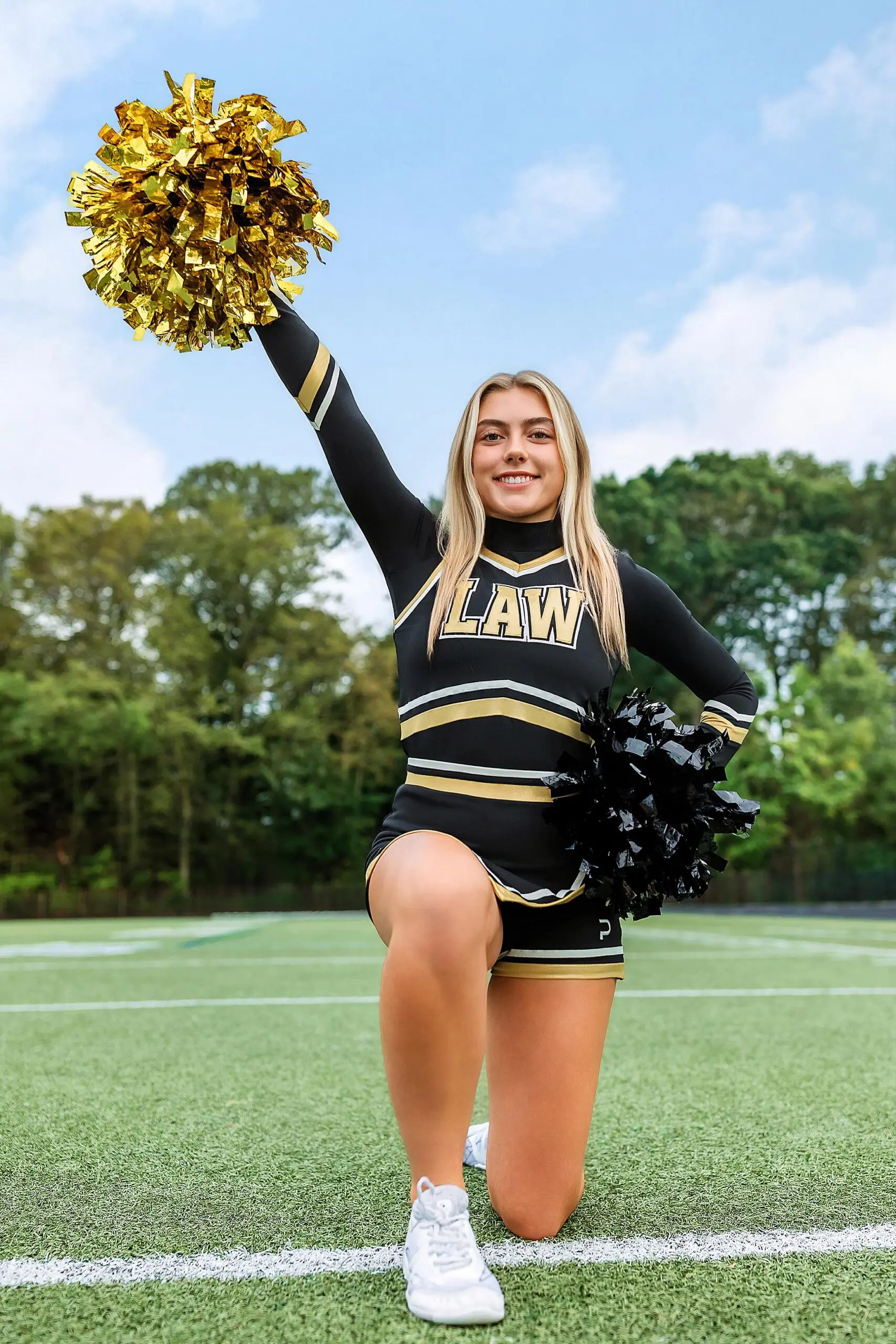 A CT senior cheerleader in a black and gold uniform kneels on one knee on the field, raising a gold pom-pom in one hand and holding a black pom-pom in the other, with trees and a blue sky in the background.