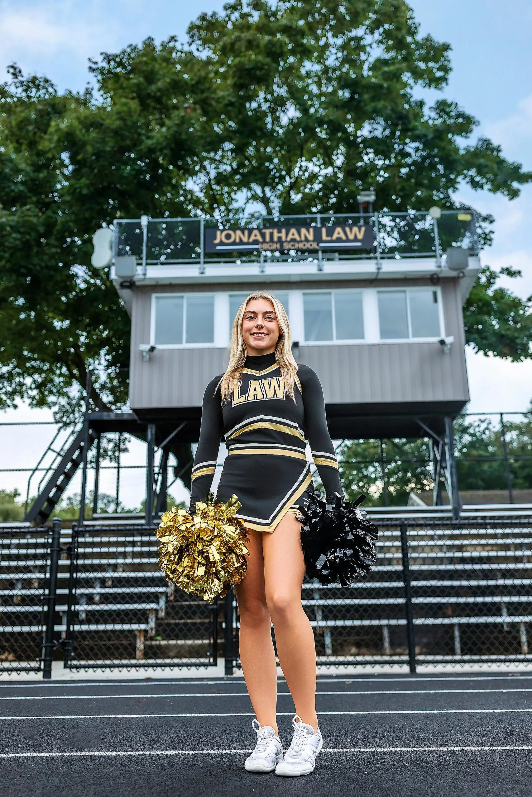 A cheerleader in a black and gold uniform holds matching pom-poms and stands on a track in front of the Jonathan Law press box, an energetic moment perfect for ct senior photos, with trees framing the background.