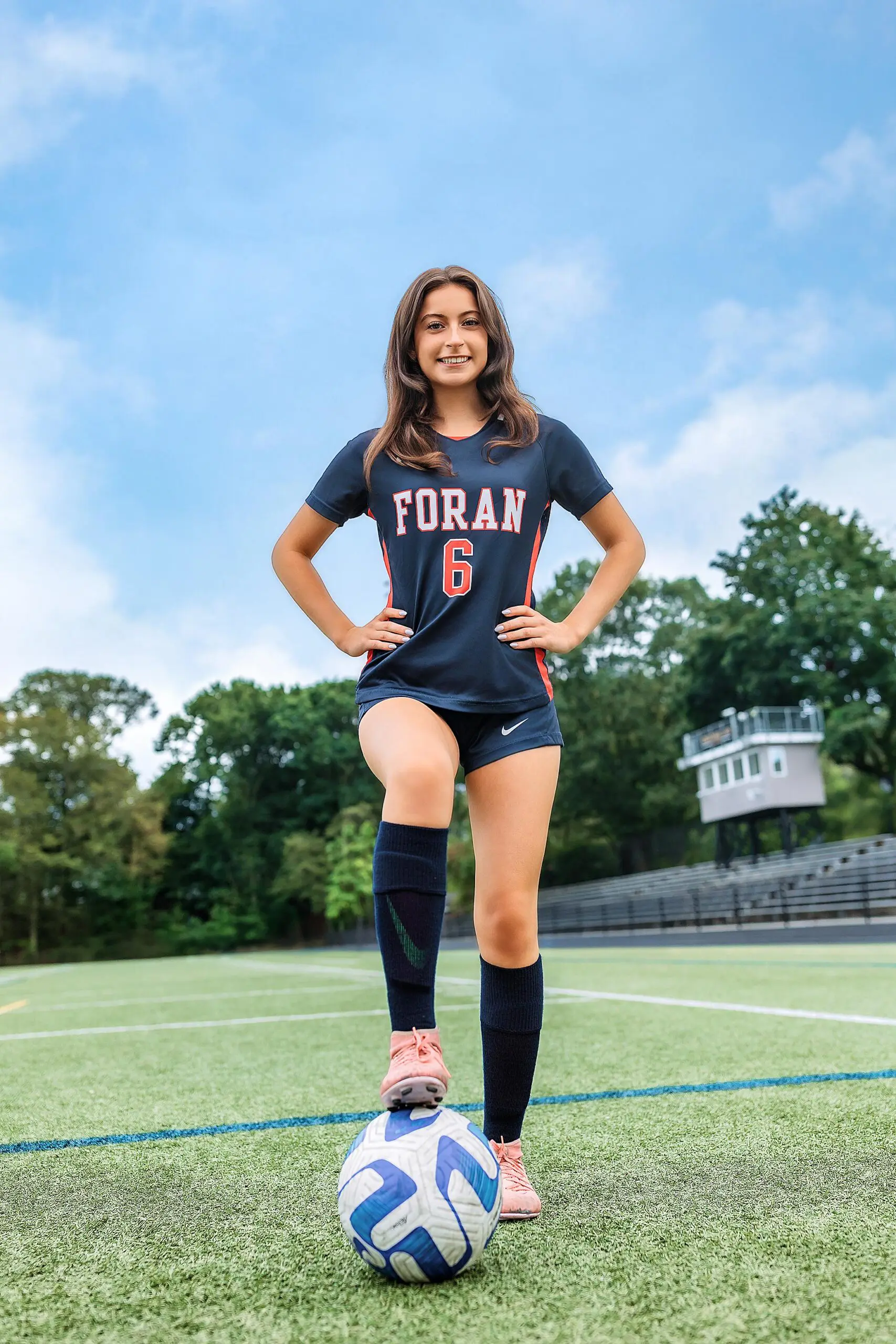 A young female soccer player in a navy FORAN jersey with number 6 stands on a green field, smiling with one foot on a blue and white ball—perfect for senior photos CT. Trees and bleachers sit in the background beneath a clear blue sky.