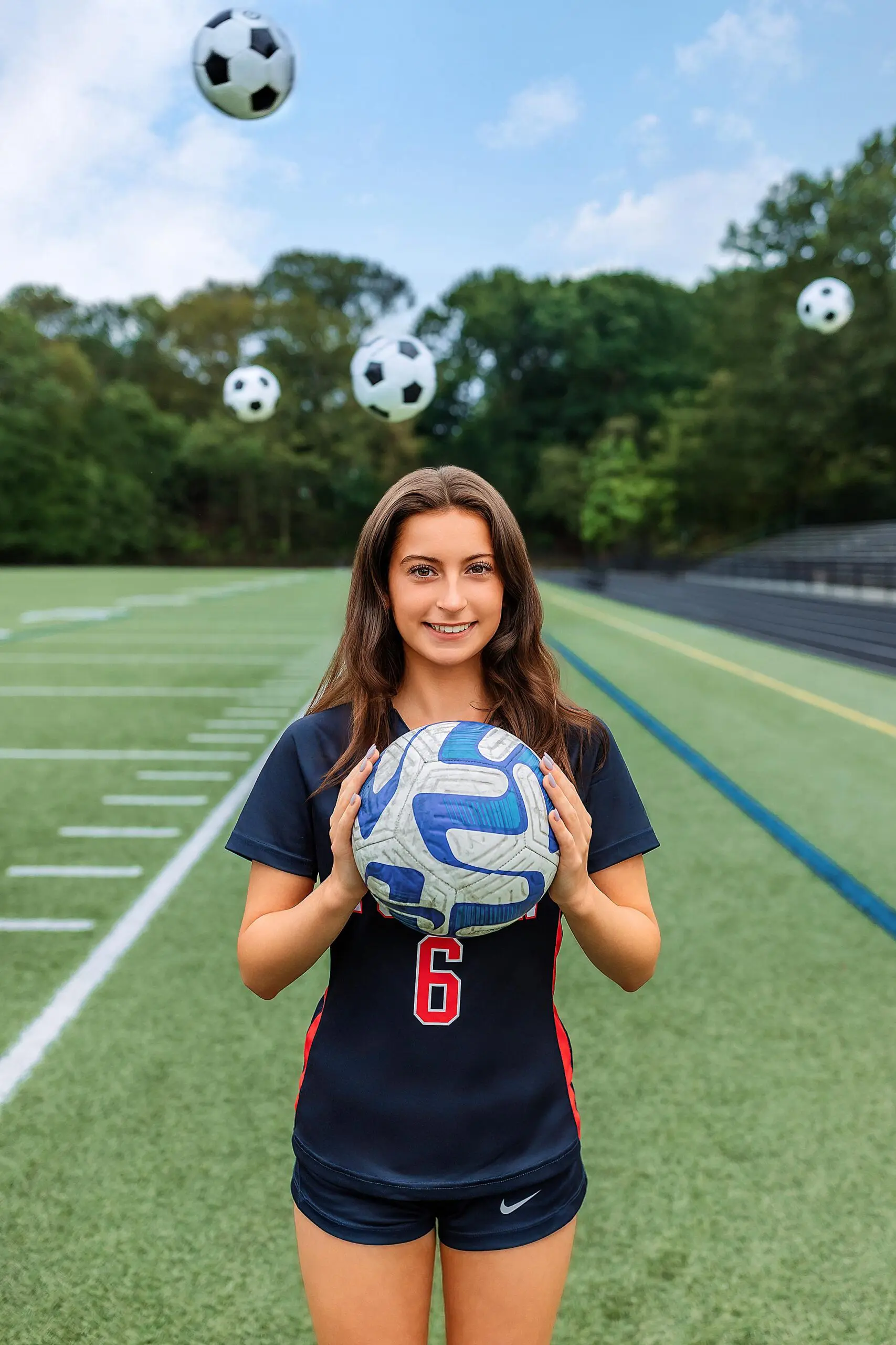 A young woman in a soccer uniform stands on a field, smiling at the camera and holding a soccer ball for her CT senior photos. Four other soccer balls float above her, with trees and bleachers visible in the background.