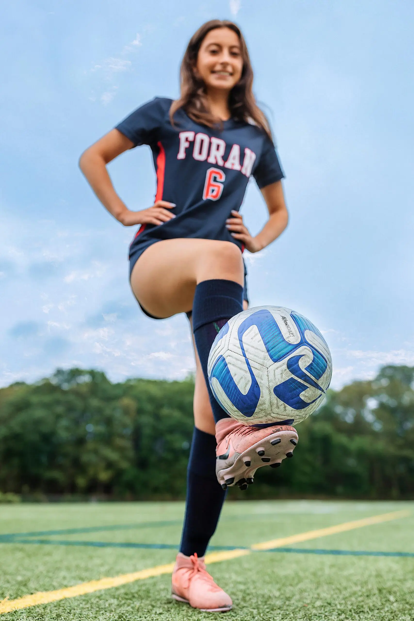 A smiling soccer player in a Foran jersey with the number 6 balances a soccer ball on her pink cleat, standing on a green outdoor field with trees in the background, perfect for vibrant CT senior portraits.