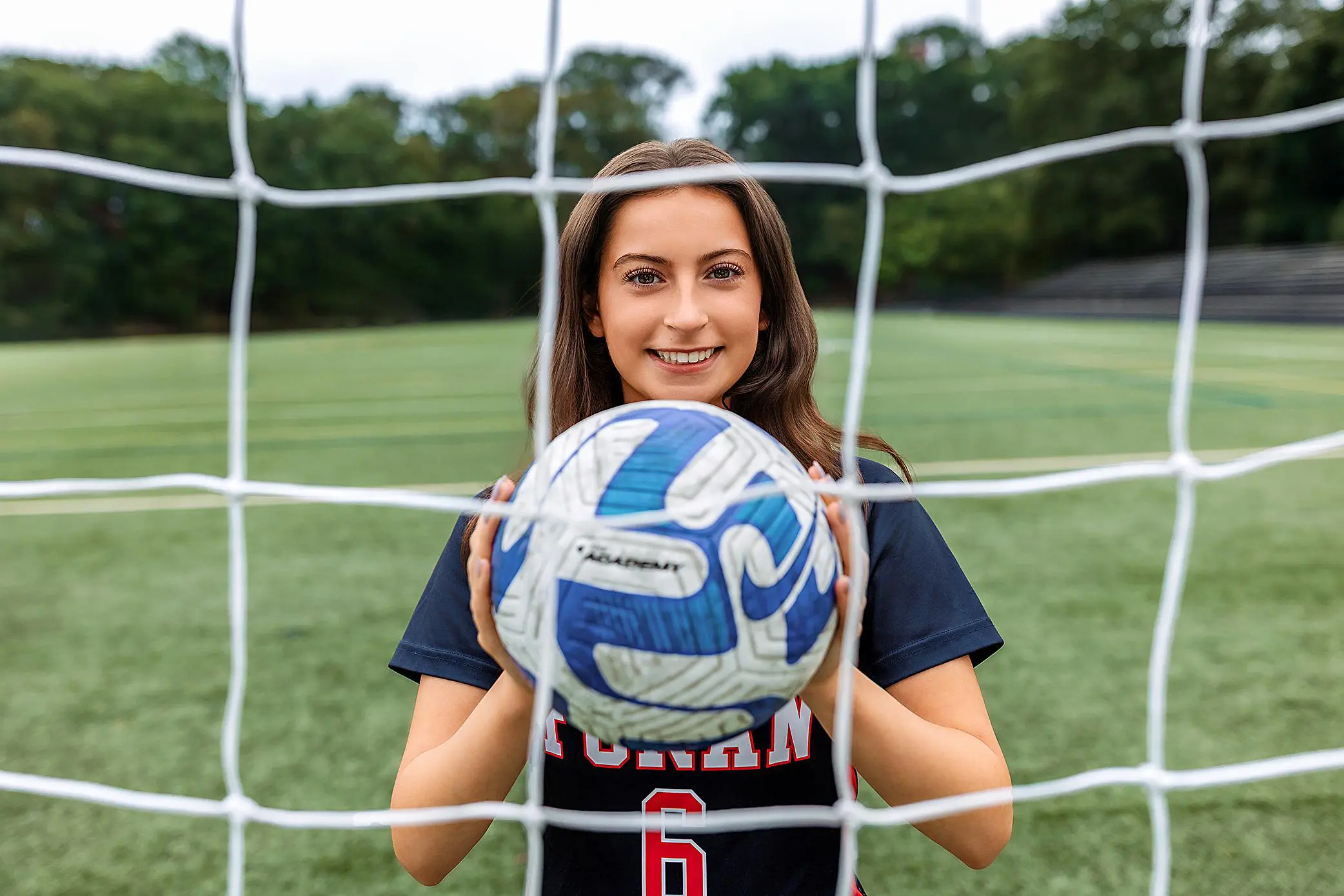 A young woman in a soccer uniform holds a blue and white soccer ball, smiling through the net on a green outdoor field during her CT senior session, with trees and bleachers in the background.