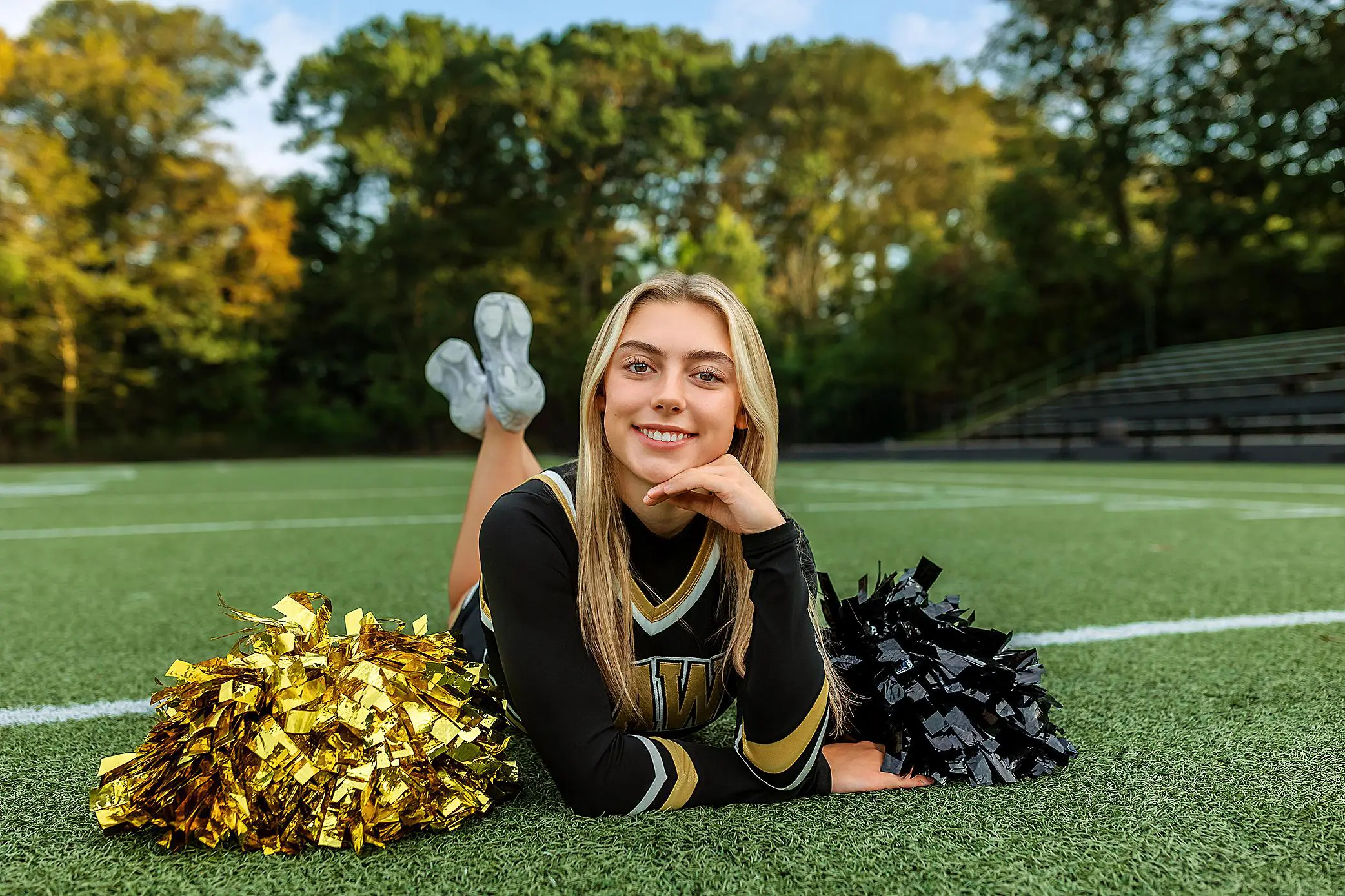 A cheerleader in a black and gold uniform lies on a football field, smiling with her chin resting on her hand. Perfect for ct senior portraits, gold and black pom-poms are in front of her, with bleachers and trees in the background.