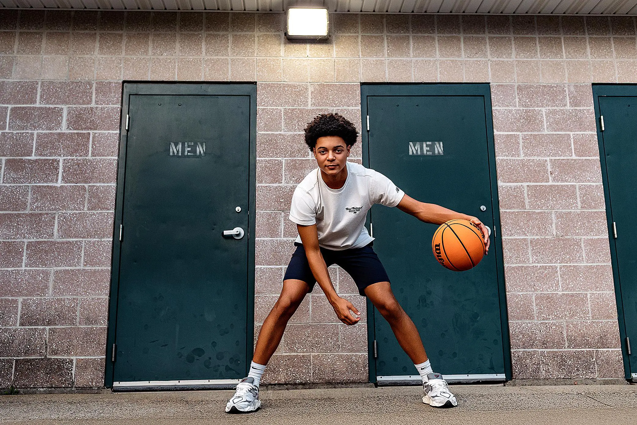 A young person in athletic clothes dribbles a basketball outside during a senior session, standing between two green doors labeled MEN on a brick building wall.