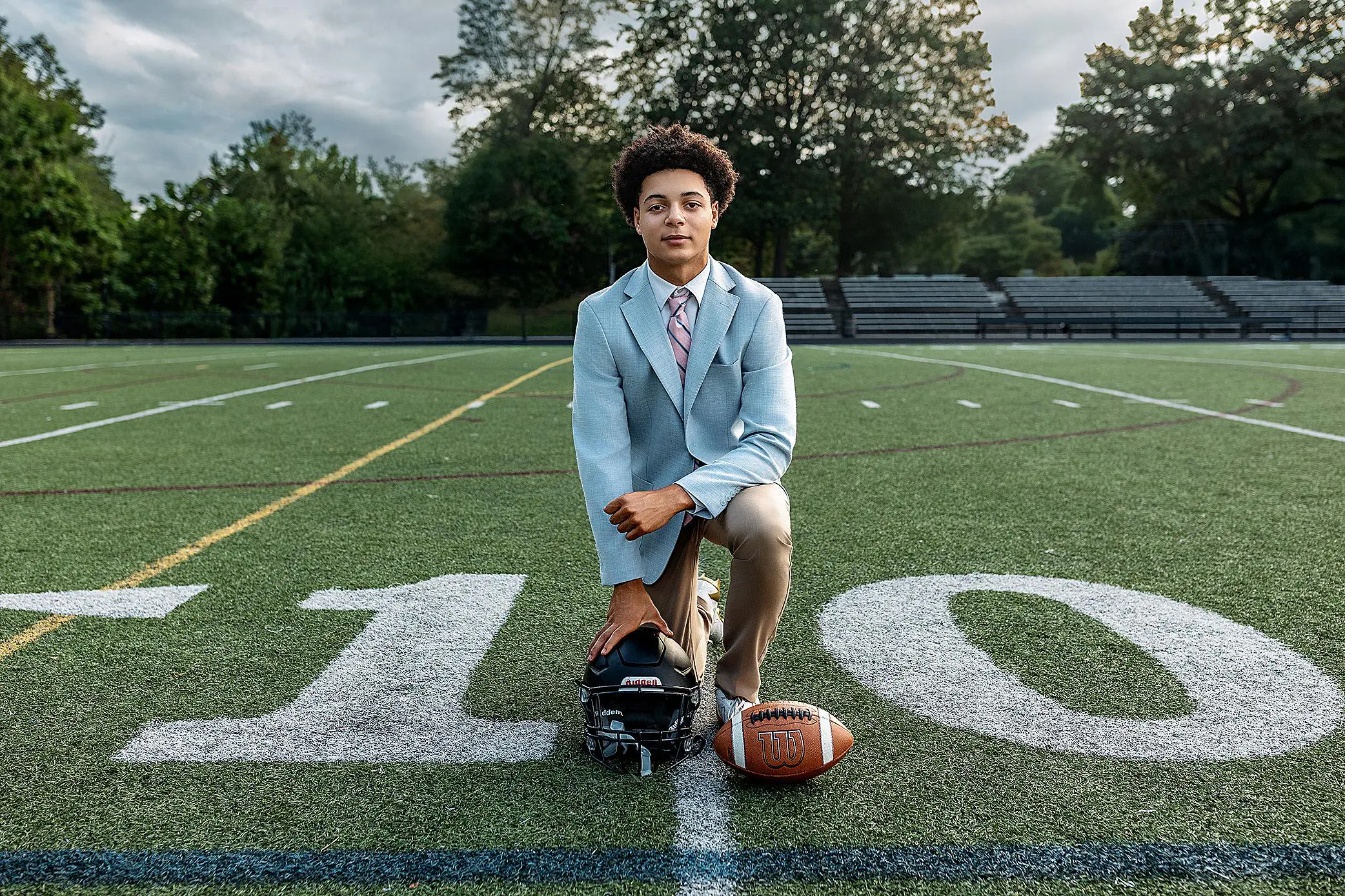 A young person in a light blue suit kneels on a football field at the 10-yard line during their CT senior session, holding a football helmet with a football nearby. Empty bleachers and trees create a serene backdrop.