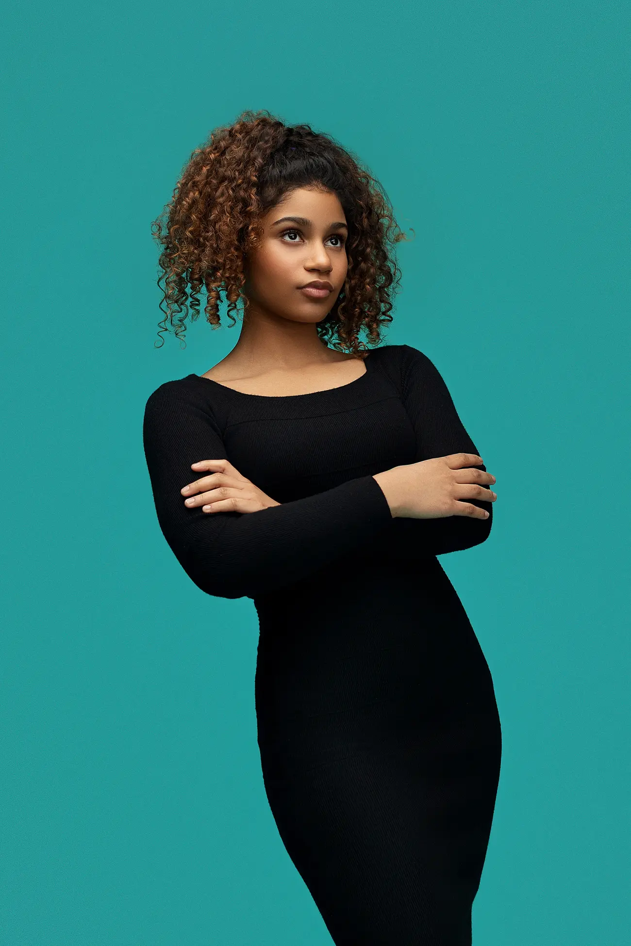 A woman with curly brown hair wearing a fitted black dress stands with arms crossed against a teal background, looking confidently to the side in this stylish connecticut-senior-portrait.