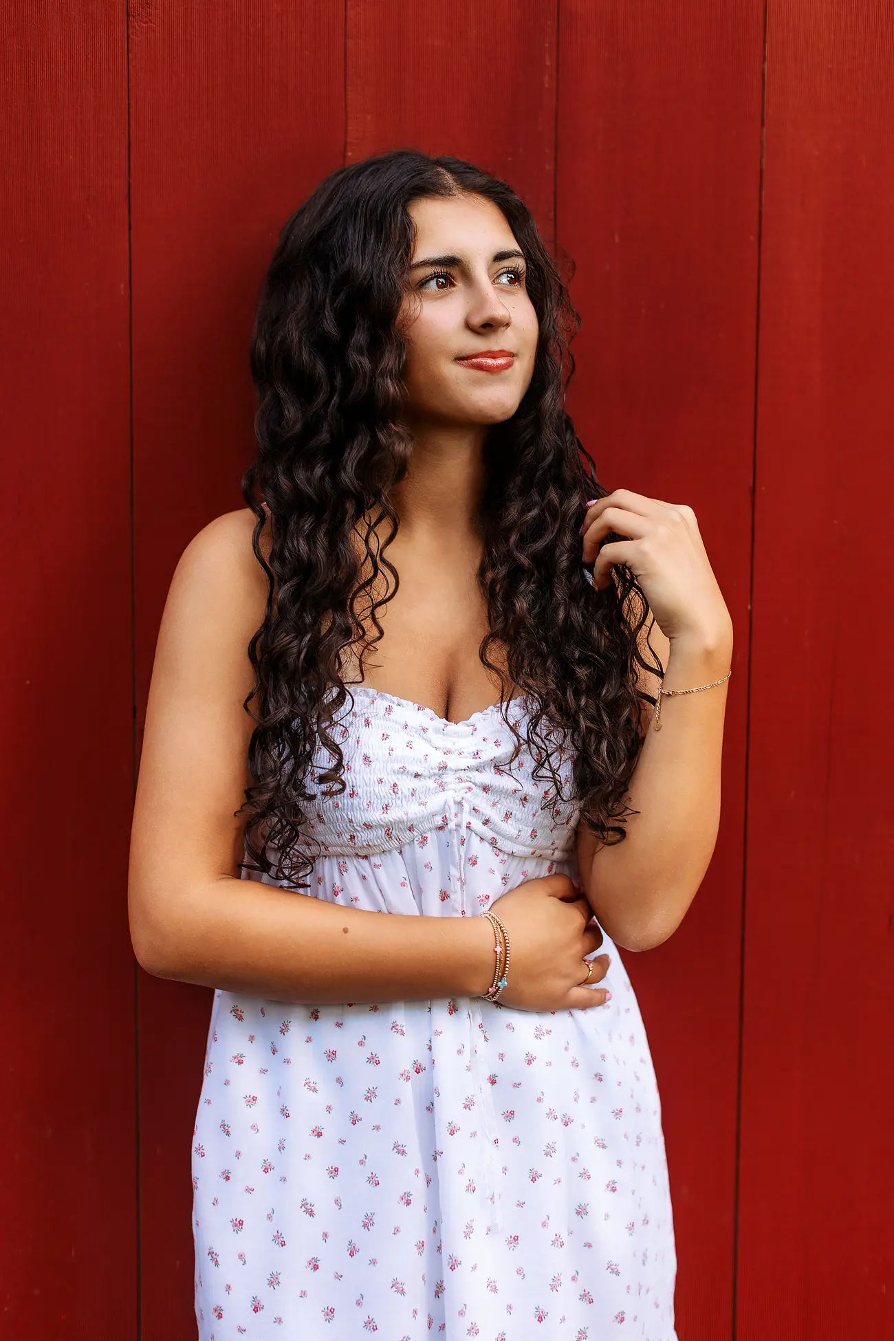 A young woman with long curly hair in a white sundress stands in front of a red wooden wall, looking to the side and gently holding her hair with one hand.