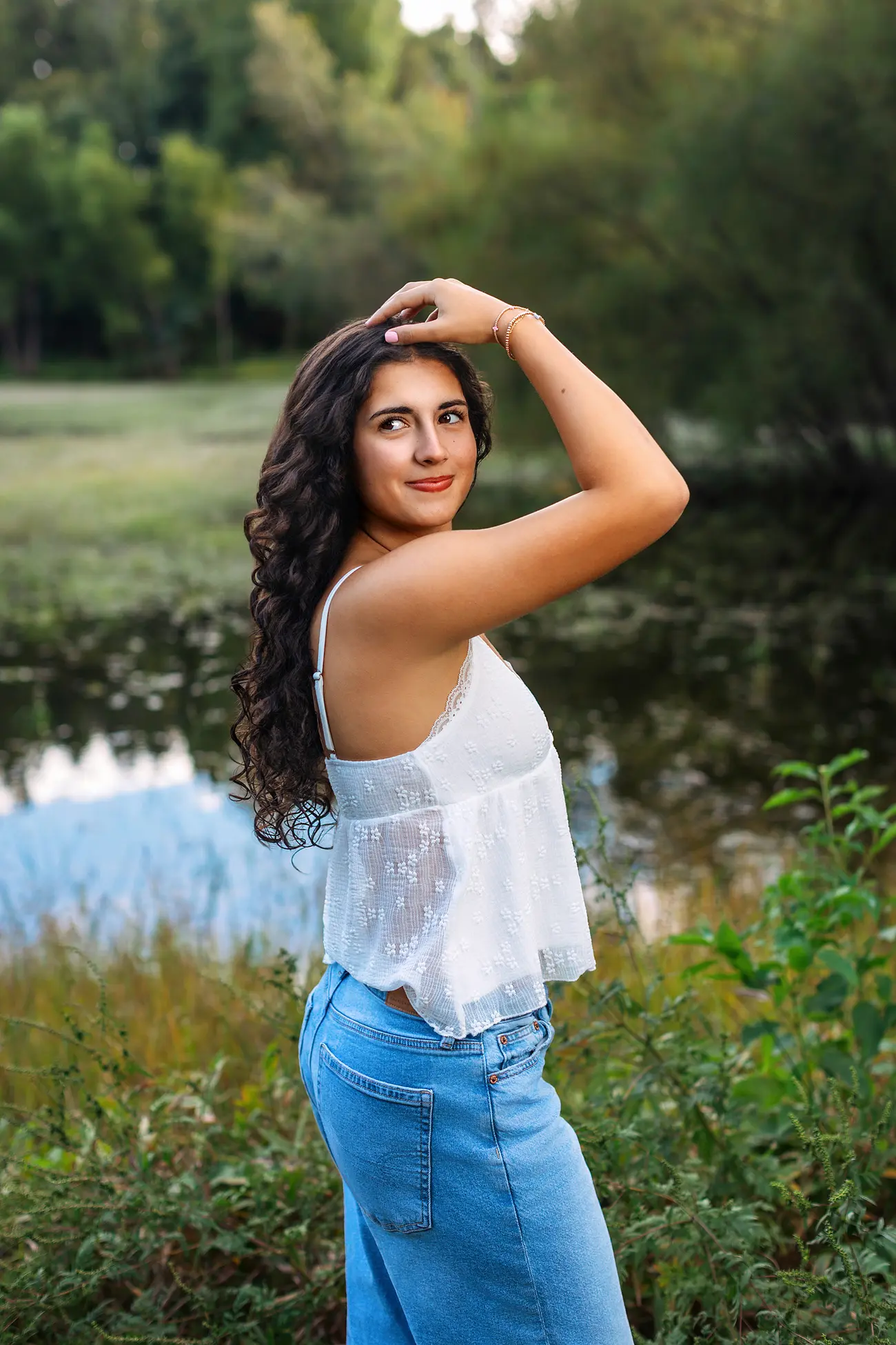 A person with long curly hair, wearing a white sleeveless top and blue jeans, poses with one arm raised by a pond in a lush, green outdoor setting.
