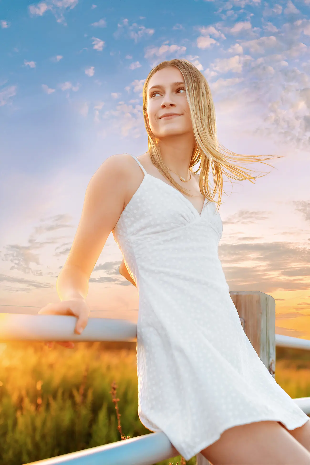 A woman in a white dress leans on a railing outdoors, smiling, with sunlight and a colorful sunset sky in the background.