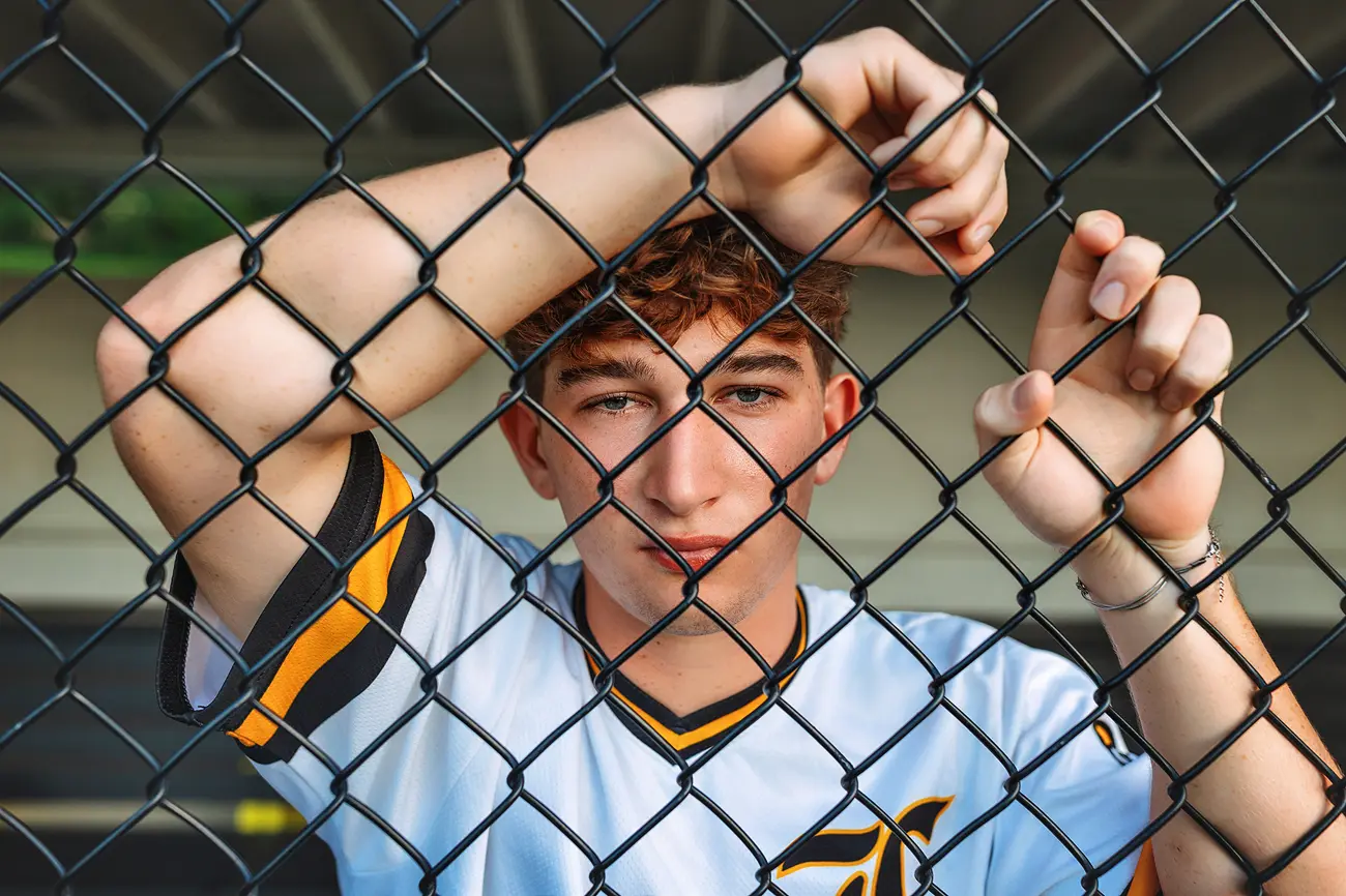 A young man in a white sports jersey stands behind a chain-link fence, holding onto the fence with both hands and looking directly at the camera.