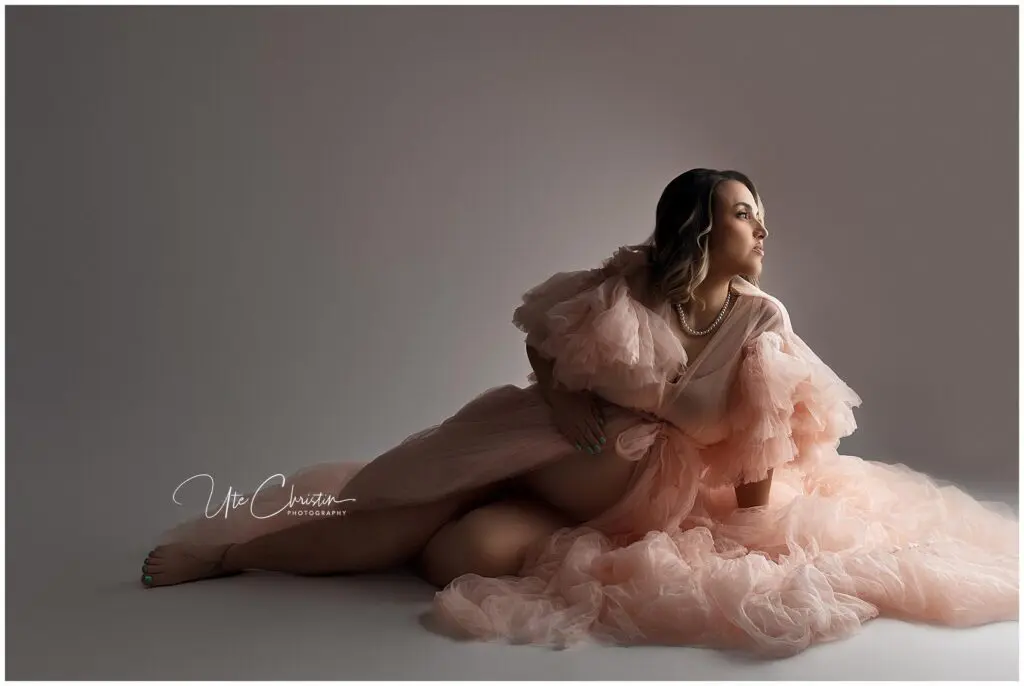 A woman in a flowing, sheer peach gown sits on the floor against a plain backdrop, looking to the side. She wears a pearl necklace and her dark hair is styled in loose waves. Soft lighting highlights her silhouette.