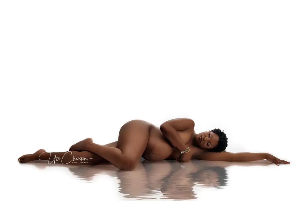 A pregnant woman lies nude on her side against a white background, one arm extended above her head and the other resting on her belly. Her image reflects softly on a glossy surface below.