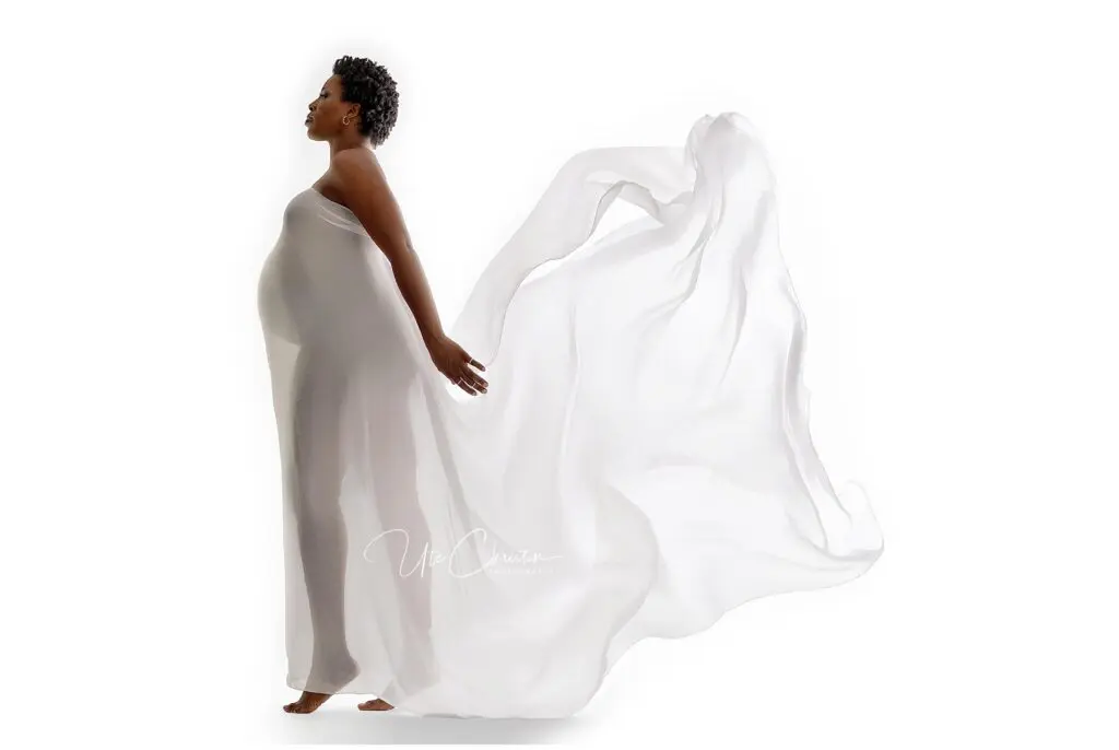 A pregnant woman stands barefoot in profile, draped in a flowing, sheer white fabric that billows behind her against a white background. She gazes forward with hands gently resting at her sides.
