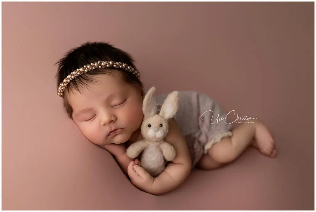 A sleeping baby with dark hair, wearing a beaded headband and a light romper, cuddles a small plush bunny while lying on a soft pink background—captured perfectly by a Milford newborn photographer.
