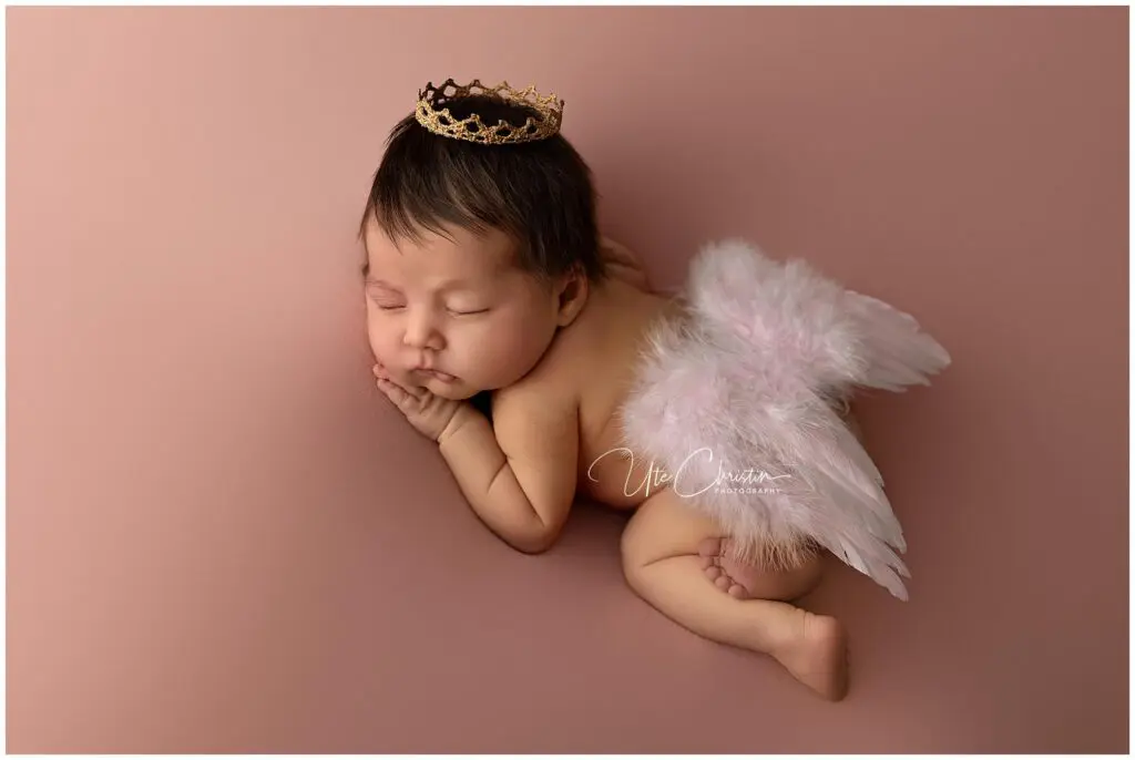 A sleeping baby lies on their stomach on a soft pink surface, wearing a small golden crown and fluffy white angel wings. Perfect for newborn photos in CT, the baby's hands are tucked under their chin, looking peaceful and serene.