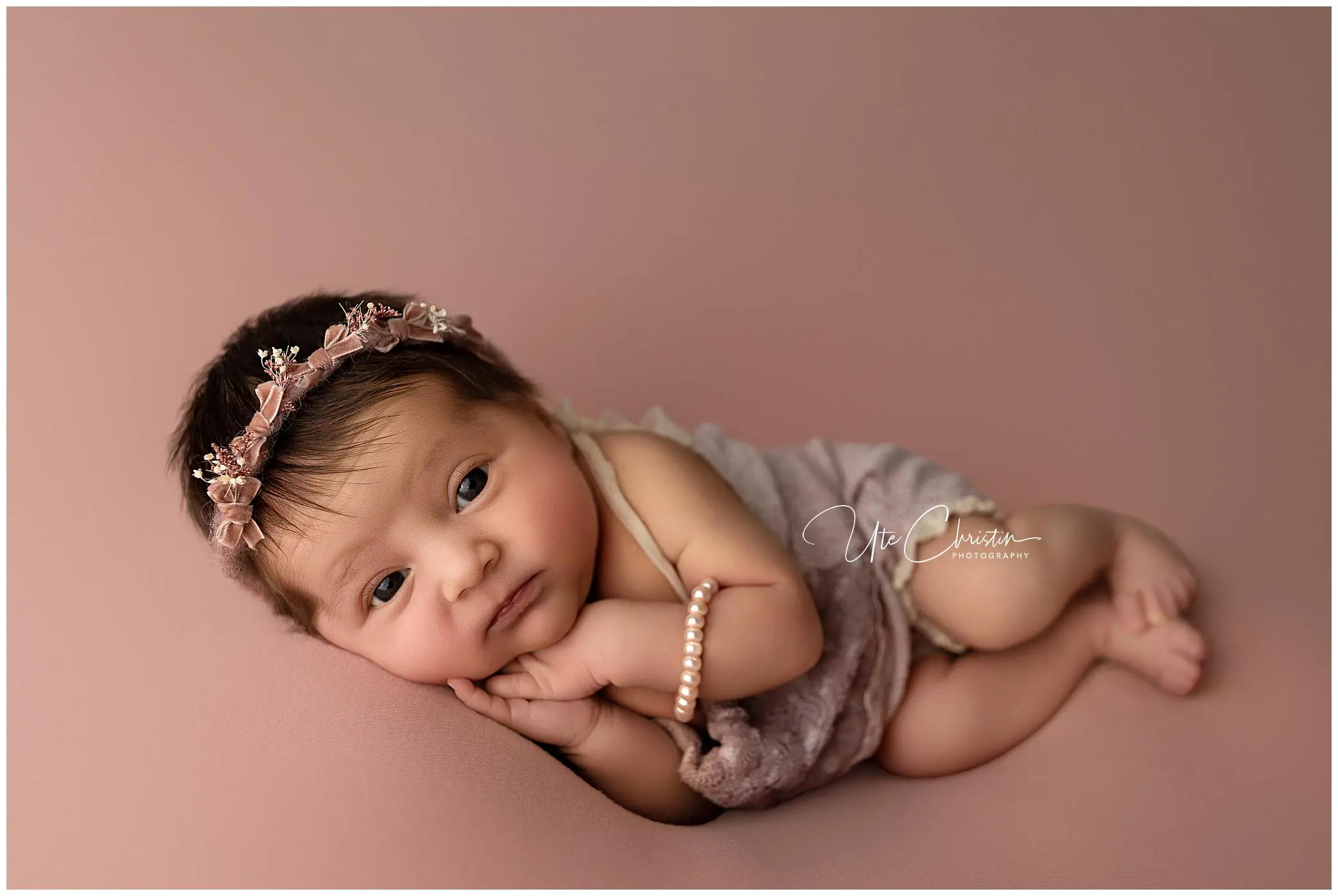 A Milford CT newborn with dark hair, wearing a floral headband and a soft mauve romper, lies on a pink blanket, resting her head on her hands and looking sweetly at the camera.
