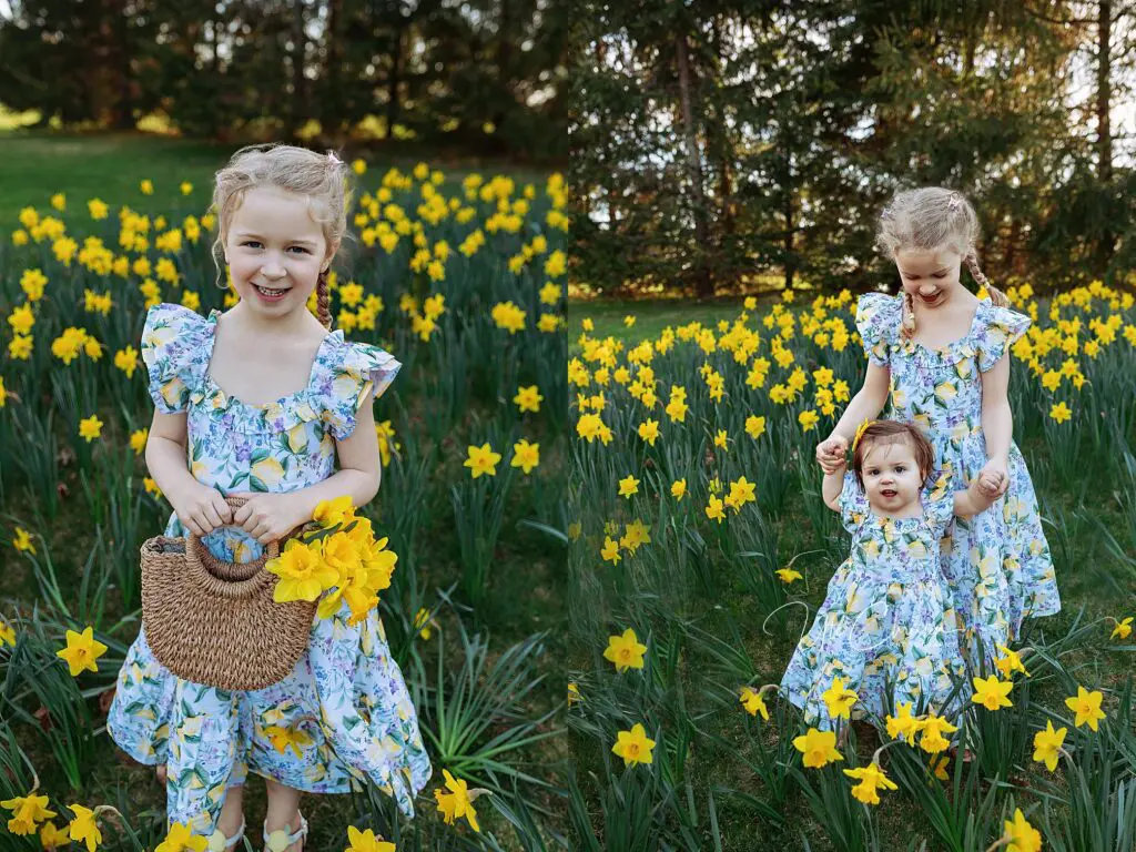 Two young girls stand in a field of yellow daffodils. Both wear matching floral dresses. The older girl holds a straw bag with flowers and smiles, while the younger holds her hand, both surrounded by blooming flowers and greenery.
