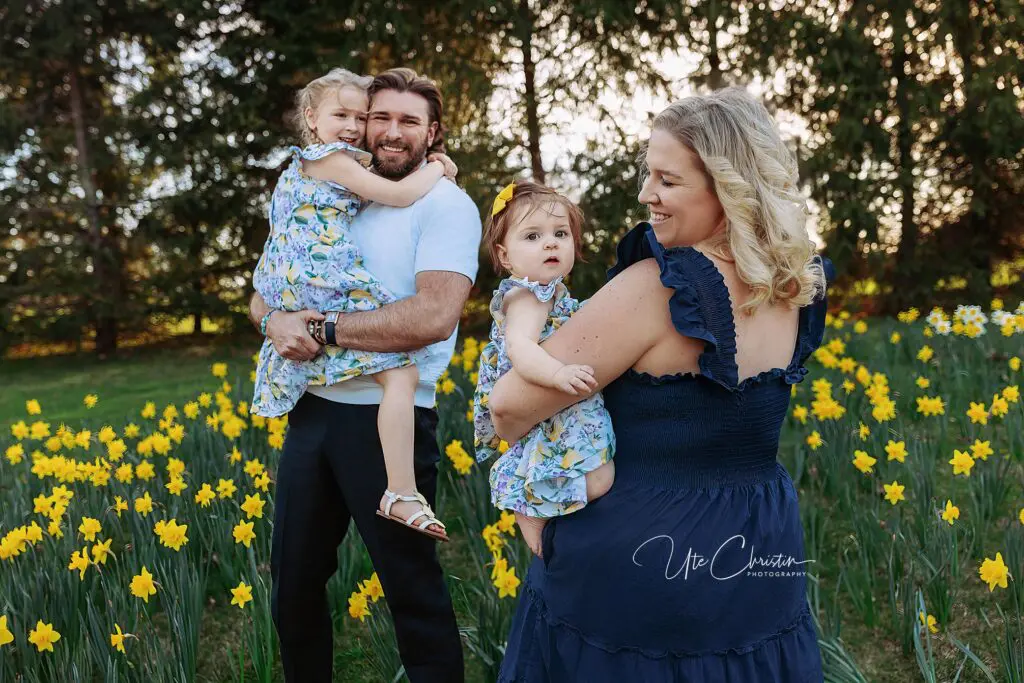 A smiling family of four stands in a field of yellow daffodils. The father holds one daughter, who hugs him and smiles. The mother holds the younger daughter, both looking toward the camera. Trees and sunlight are in the background.