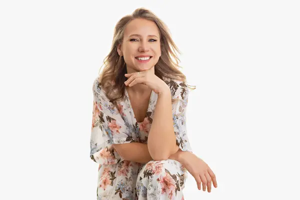 A smiling woman with blonde hair wearing a light floral dress sits with her hand resting under her chin against a plain white background. Photo of Ute-Christin Cowan a Milford Maternity Photographer.