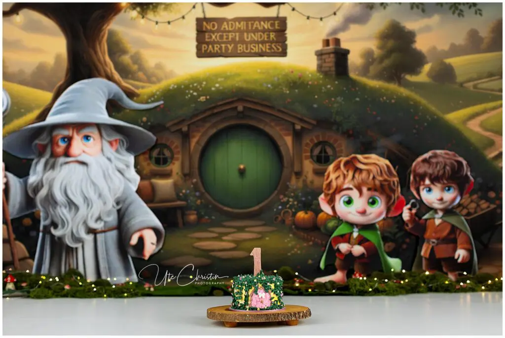 A cake with a 1 topper sits in front of a Hobbit-hole backdrop featuring cartoon versions of Gandalf and two Hobbits, with a sign reading No admittance except under party business.