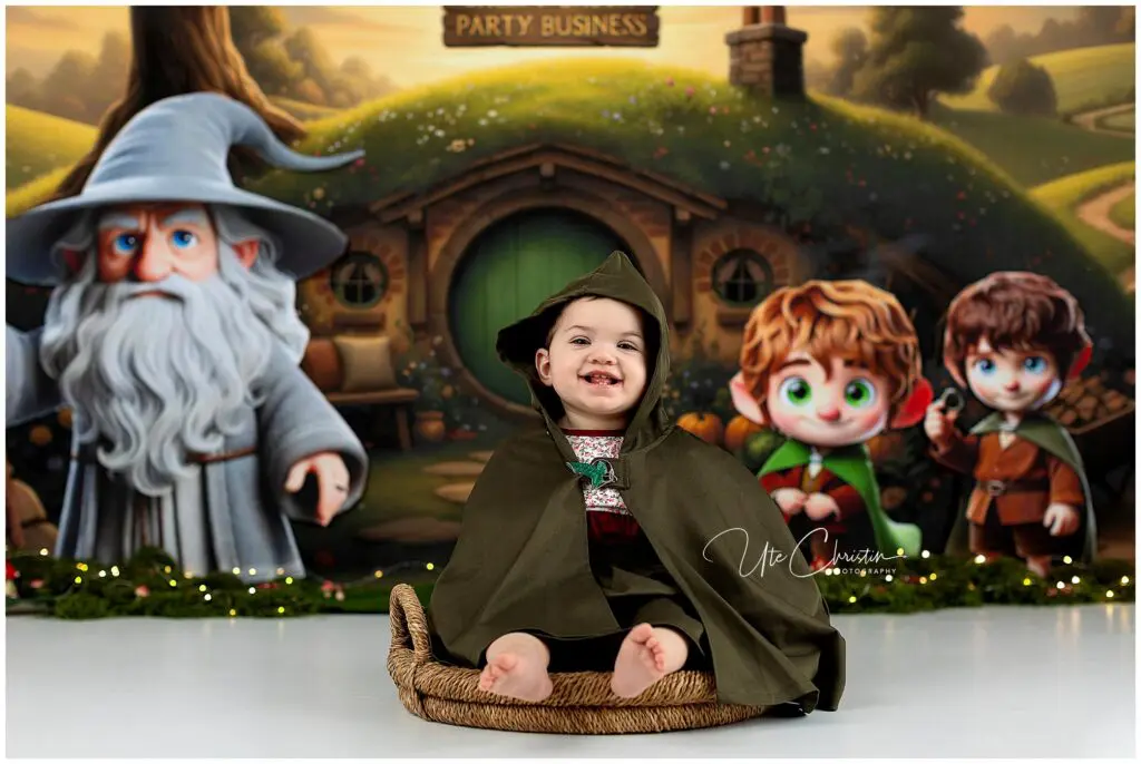 A smiling child in a green cloak sits in a basket, posing in front of a backdrop featuring cartoon versions of Gandalf, Frodo, and Sam from The Lord of the Rings, with a hobbit house and greenery in the background.