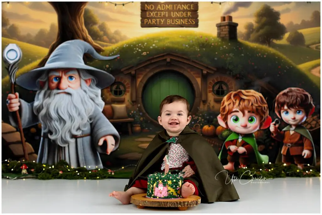 A smiling baby in a green cape sits in front of a Hobbit-themed backdrop with cartoon characters and a round door, beside a decorated cake on a wooden stand. The backdrop reads, No Admittance Except Under Party Business.