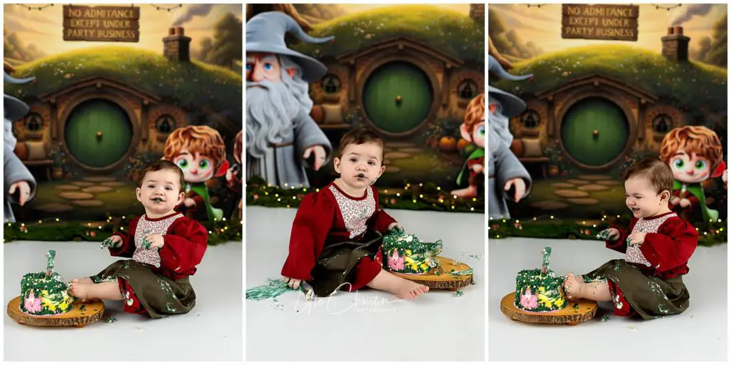 A baby in a red and green outfit sits on the floor, eating a green and yellow cake with messy hands and face, in front of a Hobbit-themed backdrop featuring characters and a round green door.