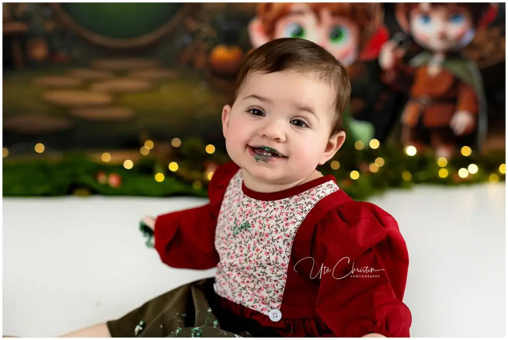 A smiling baby in a red floral dress sits on a white surface with green cake on their face and hand. There are blurred fairy lights and an illustrated fantasy backdrop behind them.