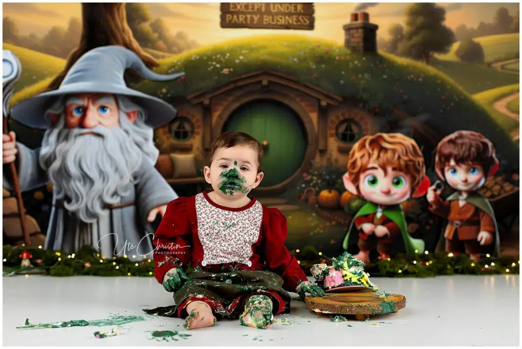 A toddler in a red outfit sits on the floor, covered in green cake and frosting, with a partially destroyed cake nearby. The background features cartoon characters resembling The Lord of the Rings in front of a hobbit house.