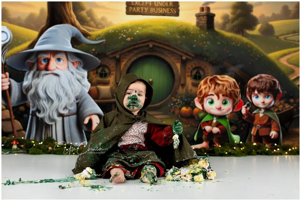 A toddler in a green Hobbit-style cloak sits on the floor covered in cake and green frosting, with a Lord of the Rings-themed backdrop featuring Gandalf and cartoon hobbits behind them.