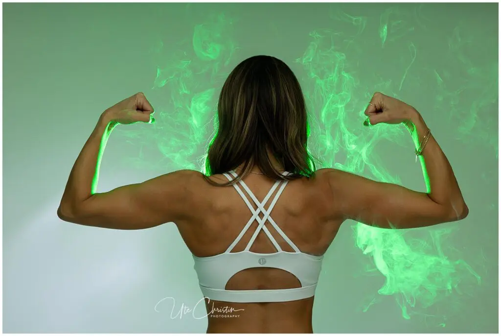 A woman in a white sports bra stands with her back to the camera, flexing her arms. Green smoke or light effects surround her muscles, highlighting her strength.