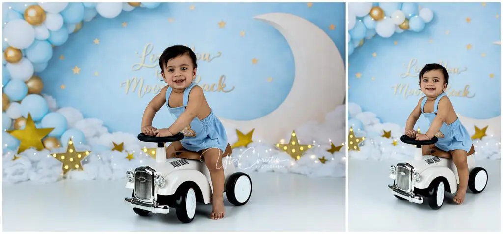 A smiling toddler in a blue dress rides a small white toy car in front of a moon-and-stars backdrop with balloons and clouds, creating a whimsical, celestial-themed scene.