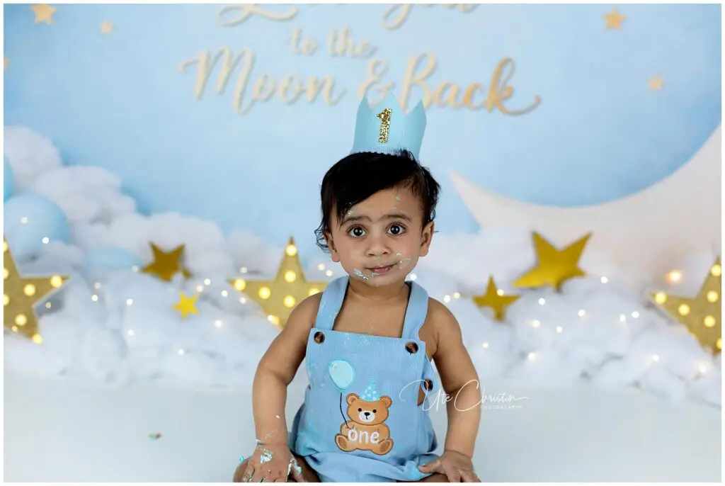 A toddler wearing a blue birthday outfit and crown with the number 1 sits in front of a Love you to the Moon & Back backdrop, surrounded by gold stars and white decorations, with cake on their face and hands.