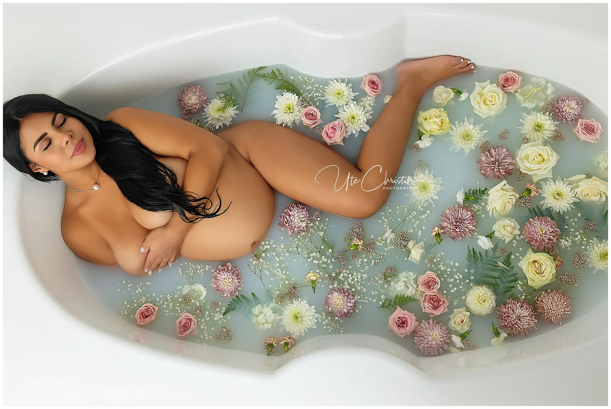 A pregnant woman lies in a milky bath filled with pink, white, and yellow flowers, gently cradling her belly and smiling with her eyes closed.
