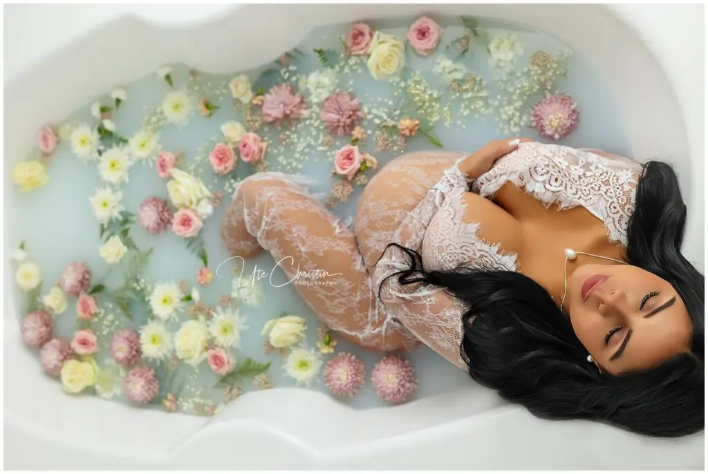 A pregnant woman in a lace robe lies in a bathtub filled with milky water and floating pink, white, and yellow flowers, gently cradling her belly and looking serene.