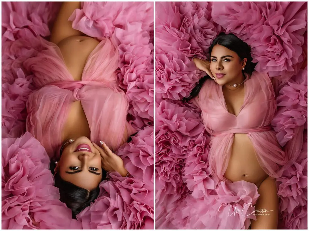 A pregnant woman in a sheer pink gown lies on ruffled pink fabric, cradling her belly and smiling at the camera in an elegant maternity photoshoot.