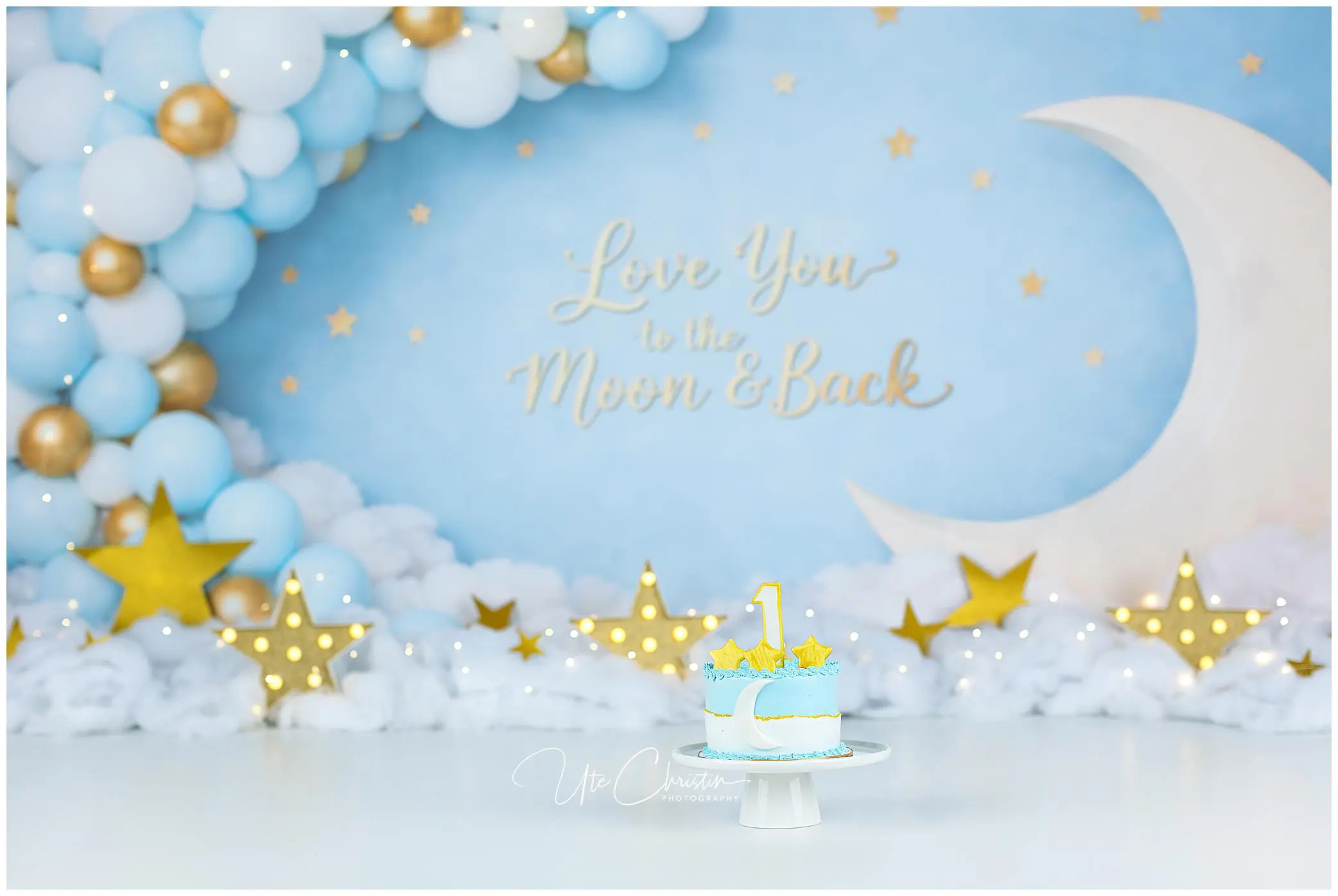 A blue and white cake with a yellow number one topper sits on a stand in front of a Love you to the moon & back backdrop, decorated with balloons, gold stars, and a crescent moon.