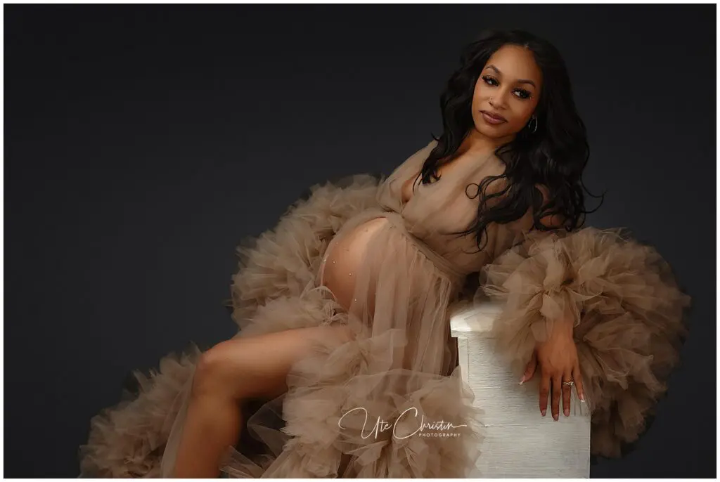 A pregnant woman in a sheer, ruffled, light brown gown poses gracefully on a white stool against a dark background, smiling softly at the camera with one hand resting on her baby bump.