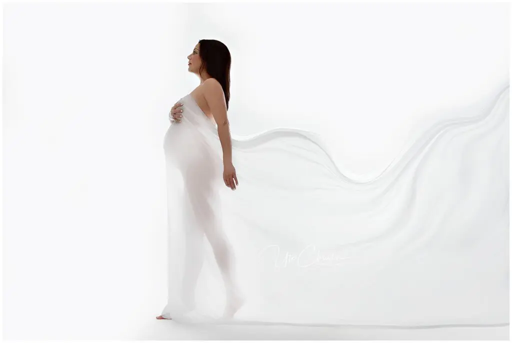 A pregnant woman stands in profile, draped in sheer white fabric that flows behind her, set against a plain white background. She cradles her belly and looks forward with a serene expression.