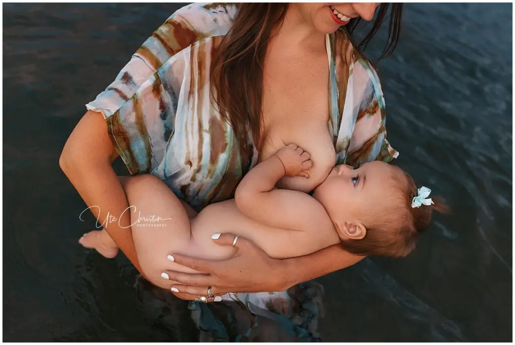 A woman in a colorful, flowy top breastfeeds her baby with a blue bow while standing in water. Both appear content and relaxed, sharing an intimate moment perfect for a newborn photographer CT to capture.
