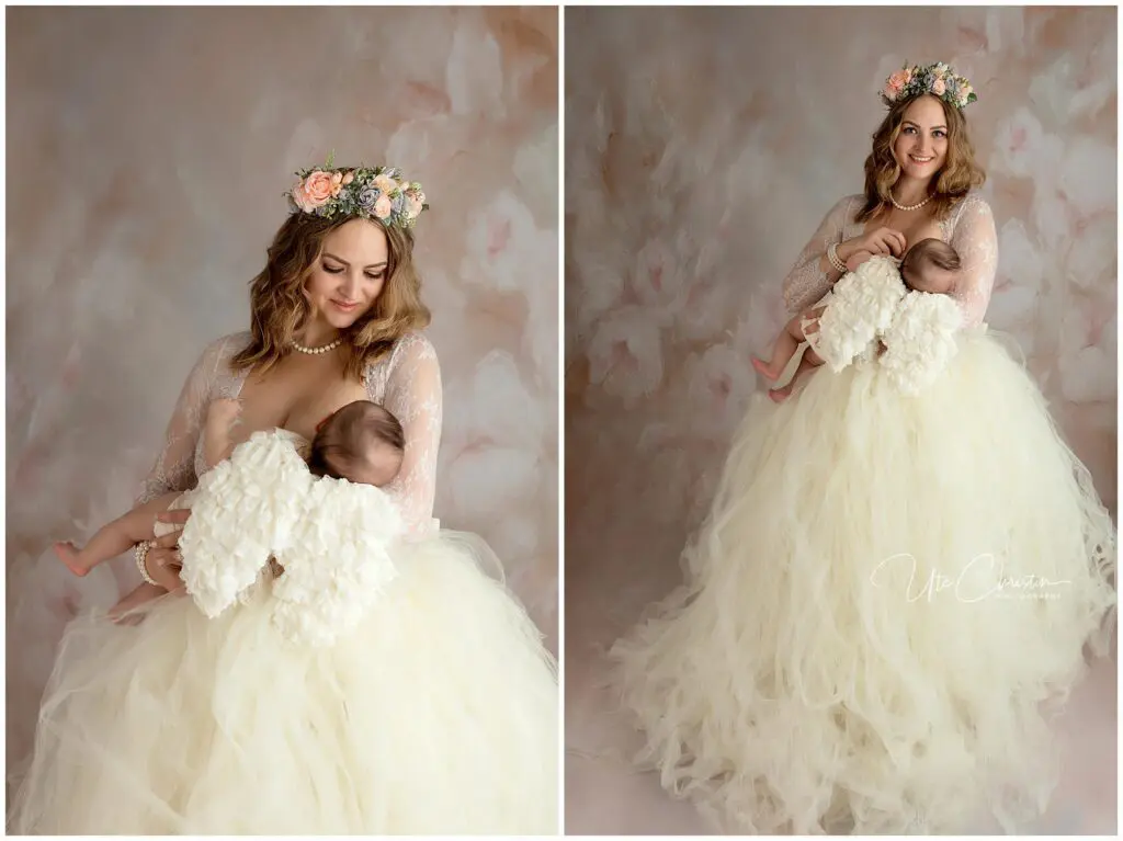 A smiling woman in a long, flowing cream gown and floral crown lovingly cradles a baby wrapped in a soft white blanket, standing against a soft, pastel background.