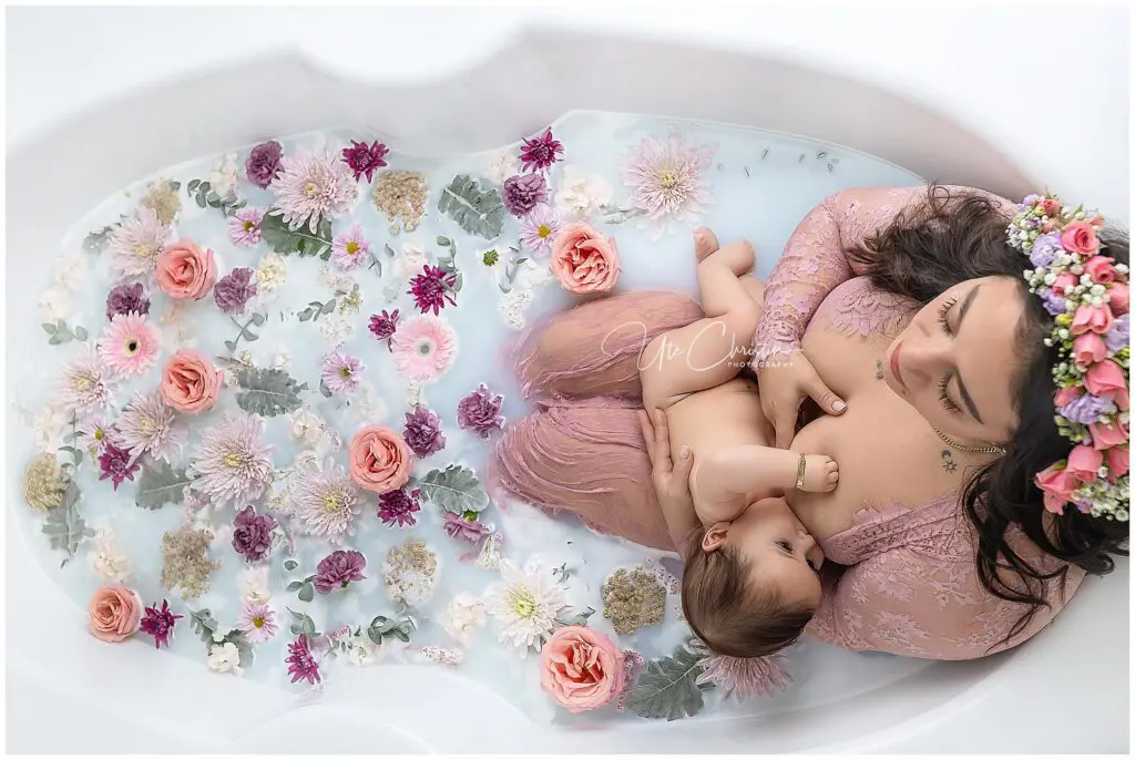A mother in a pink lace dress and floral crown breastfeeds her baby in a milky bathtub adorned with colorful flowers, capturing the serenity of mastering the art of breastfeeding in Milford.