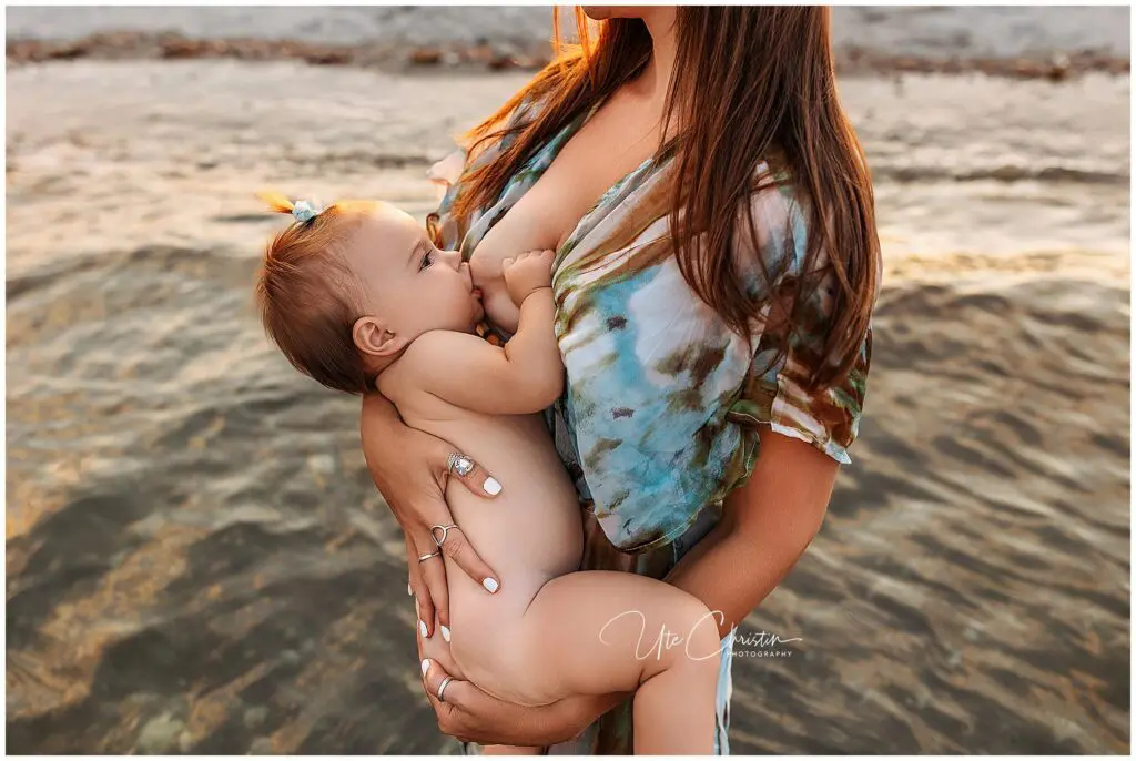 A woman stands in shallow water breastfeeding a baby with a small bow in their hair. The woman wears a patterned dress, and the background shows gentle waves and sandy shore.